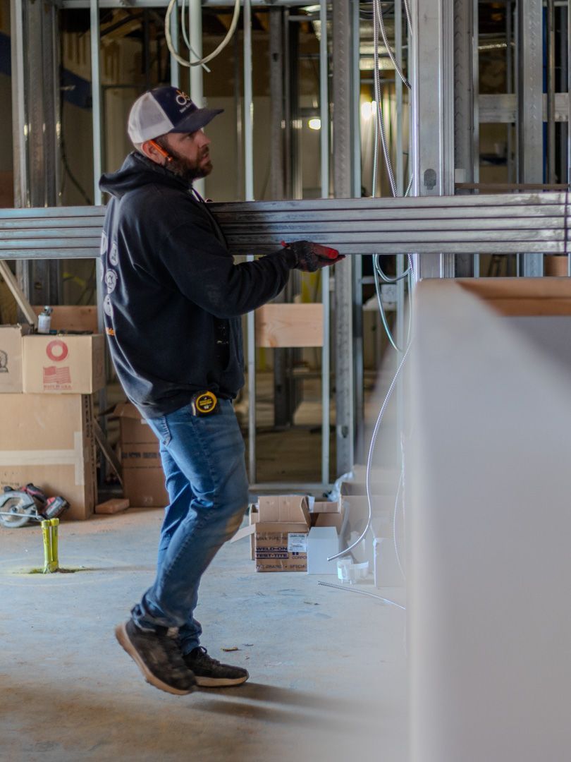 A worker in a dark hoodie and cap carries a stack of metal framing studs through an indoor construction site.