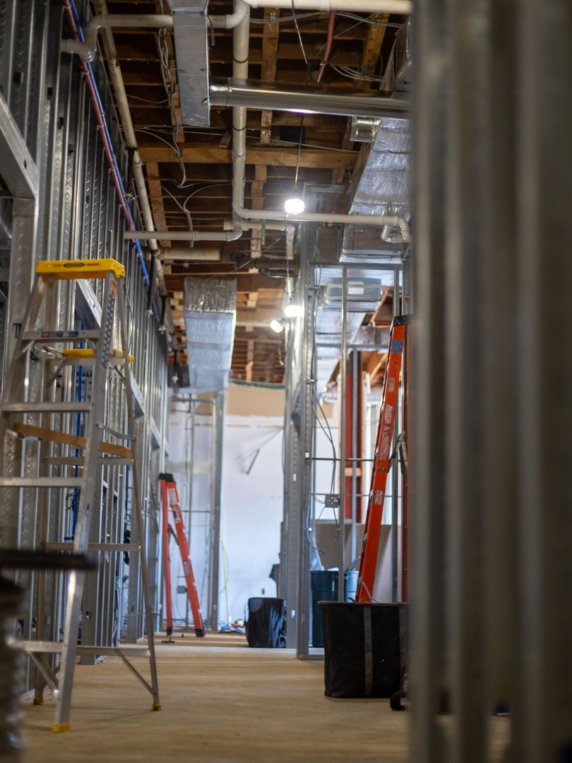 An interior view of a building under construction, featuring metal framing, exposed ceiling pipes, and ladders.