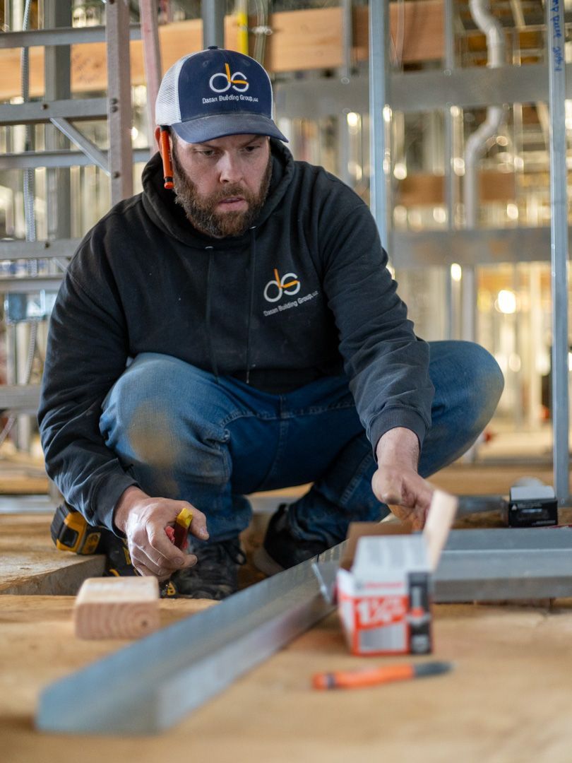 A person wearing a dark hoodie and logo cap crouching on a construction site, holding a tool while working on metal framing.