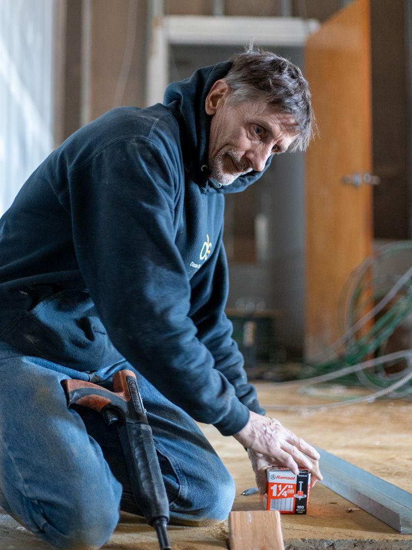 A person in a dark hoodie kneels on a construction site floor, holding a small box and a tool, working on home repairs.
