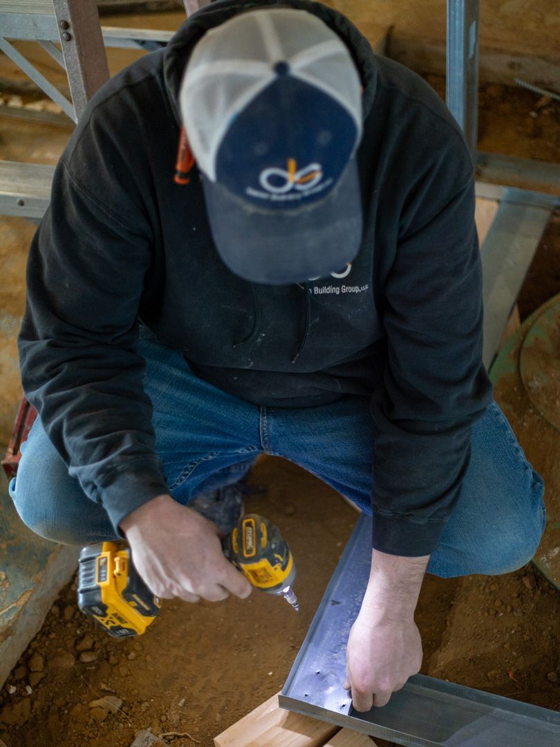 A person wearing a hat and black sweatshirt uses a yellow drill to attach a metal beam on a construction site.