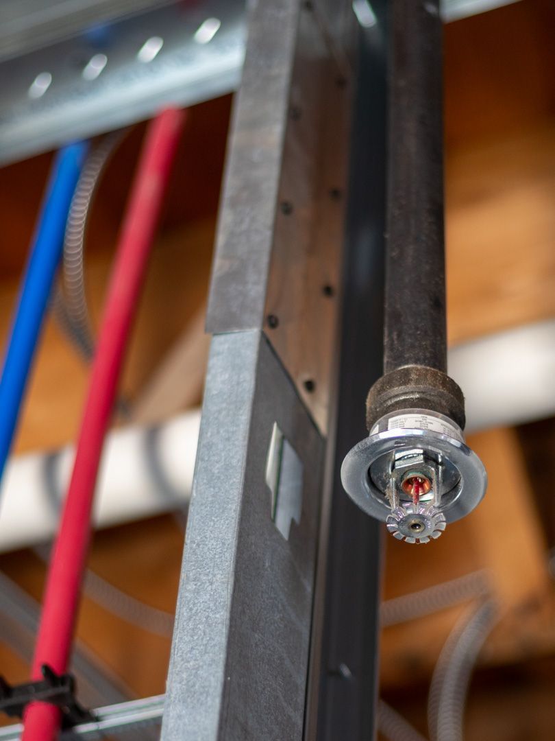 A fire sprinkler head mounted on a dark pipe, positioned next to a metal stud and red and blue plastic pipes in a ceiling.