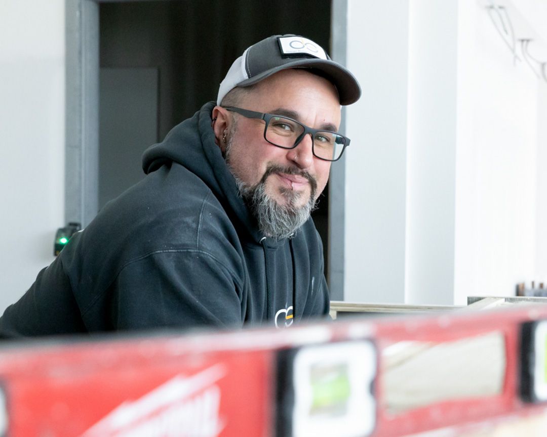 A person wearing a cap, glasses, and a dark hoodie smiles behind a red level tool in a brightly lit indoor space.