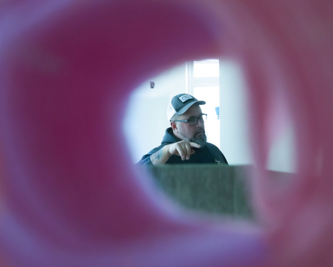 A person wearing a cap and glasses gesturing while speaking, viewed through a soft, out-of-focus pink circular frame.