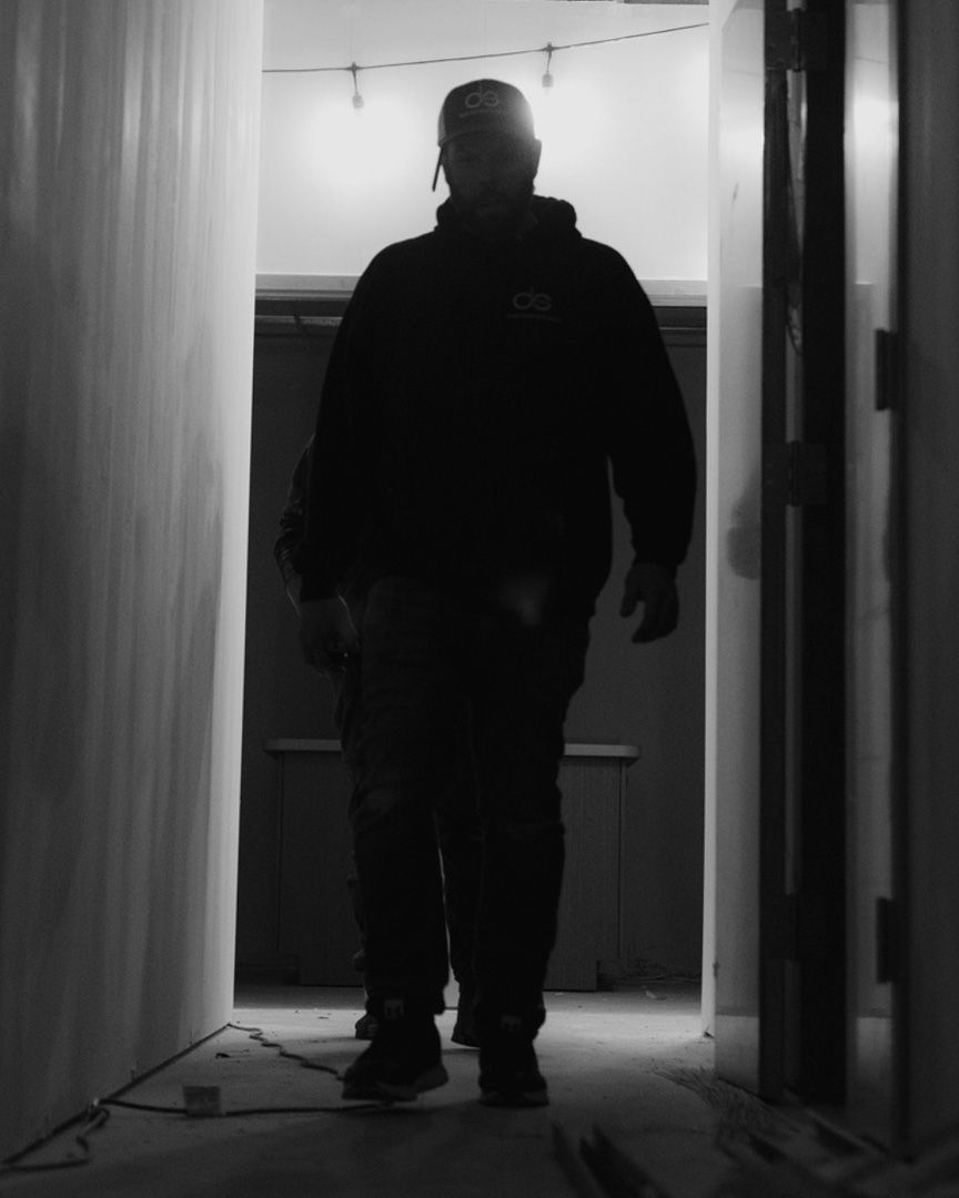 A person wearing a hooded sweatshirt walks toward the camera through a dimly lit, narrow corridor.