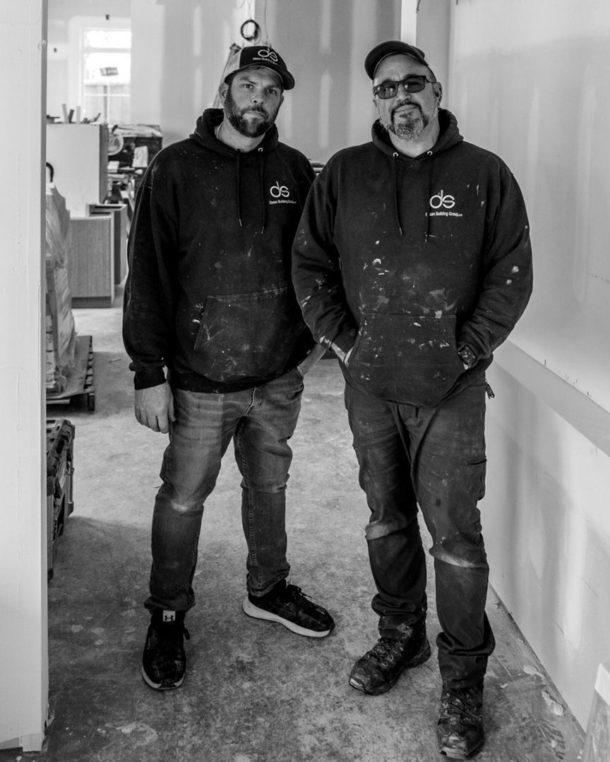 Two people in paint-splattered hoodies stand side-by-side in a room under construction.