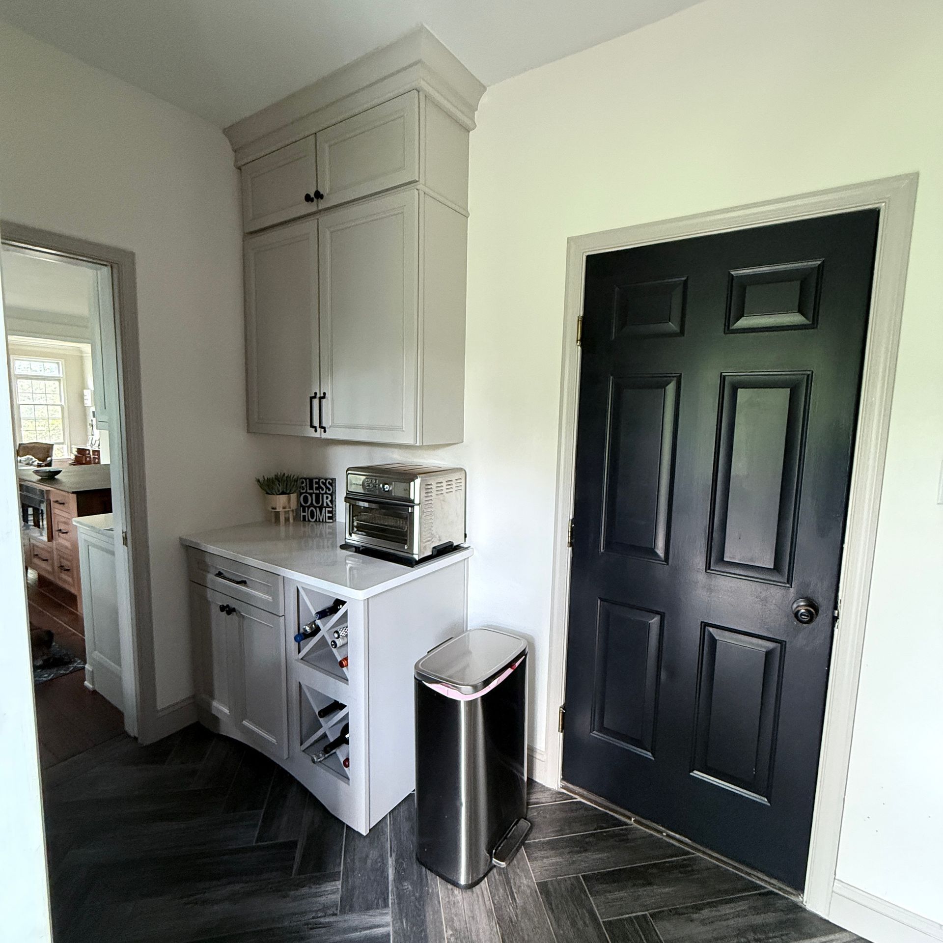 A light grey cabinet and counter with a toaster, wine rack, and metal trash bin next to a black door in a home entryway.