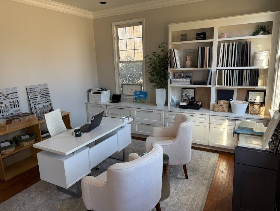 A white desk, two light chairs, and a large built-in bookshelf fill a brightly lit office with neutral-toned walls.