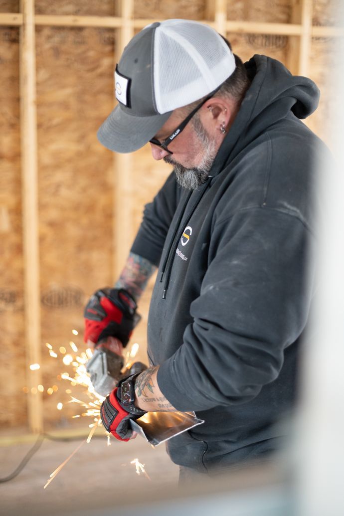 A person wearing a baseball cap and work gloves uses a handheld grinder on metal, creating sparks inside a wood-framed room.