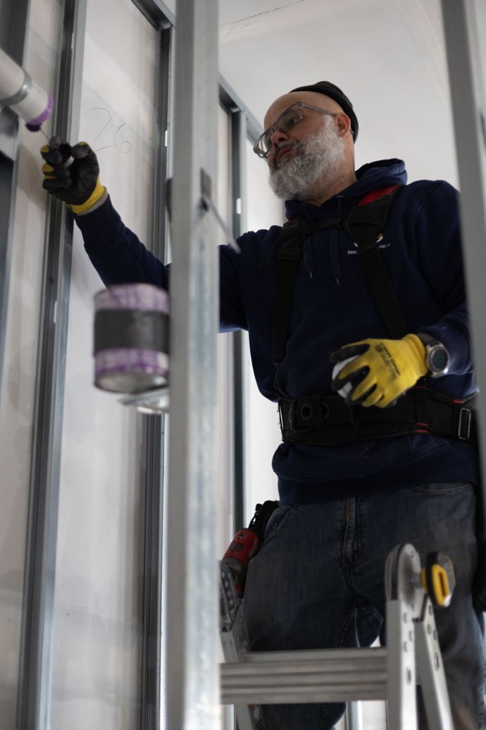 A worker wearing safety gloves stands on a ladder, installing piping within a metal frame structure for a wall.