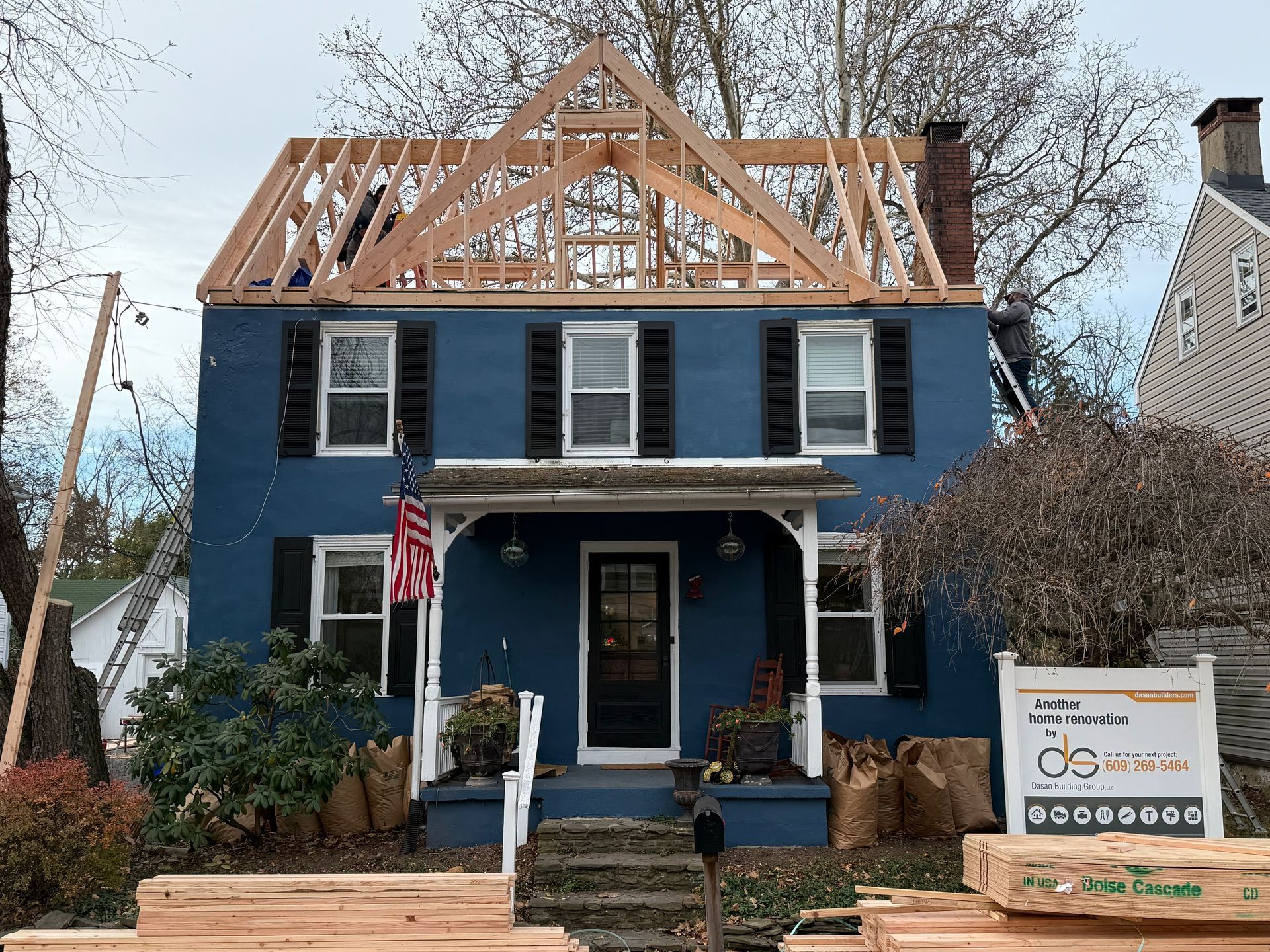 A blue two-story house undergoing roof renovations with exposed wooden framing and a construction sign in the front yard.