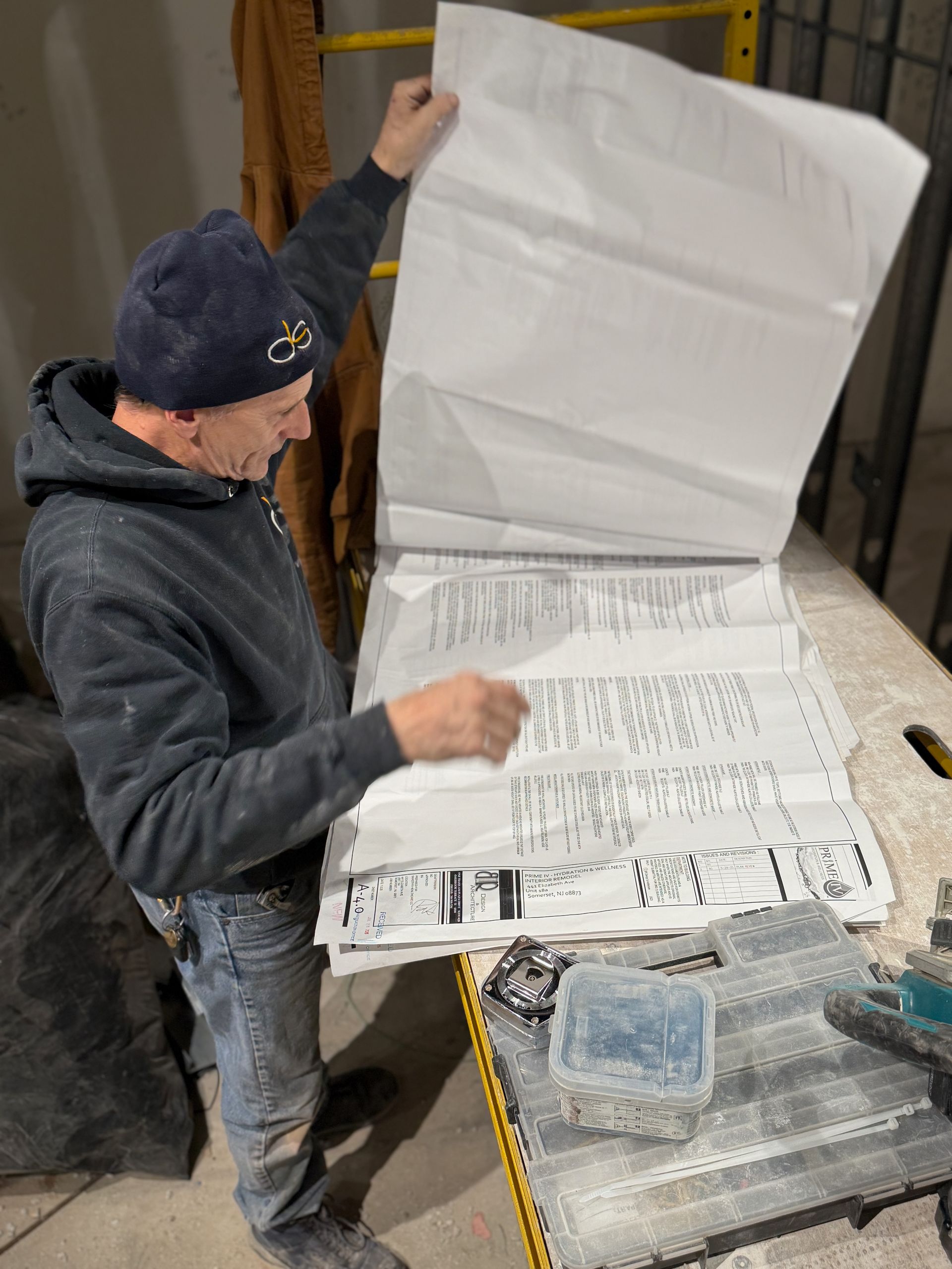 A worker in a dark hoodie and beanie reviews a large, unfolded blueprint on a workbench in a workspace.