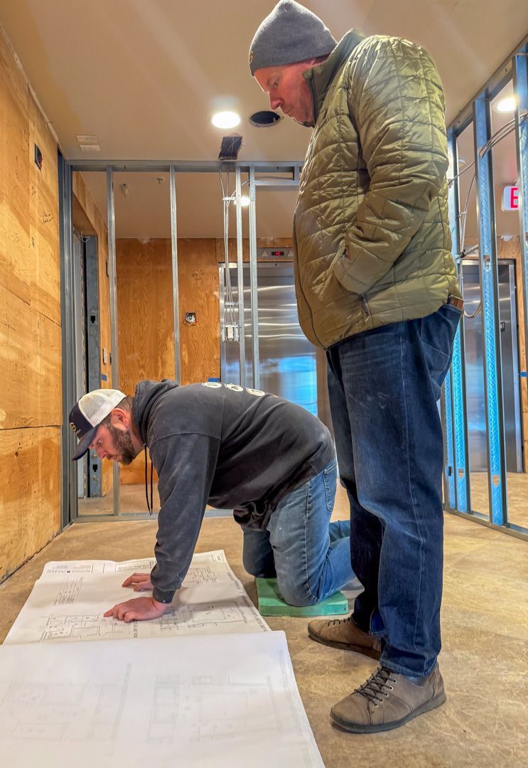 Two workers in a construction site looking at blueprints spread out on the floor.