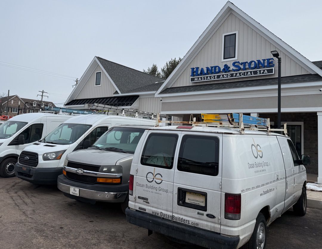 Three white work vans parked in front of a Hand & Stone Massage and Facial Spa building on an overcast day.
