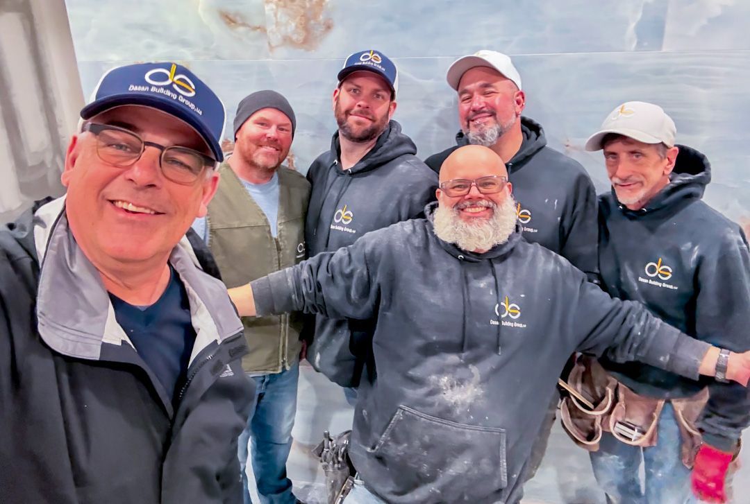 A group of six smiling people wearing branded work hoodies and hats pose together indoors, likely at a construction site.