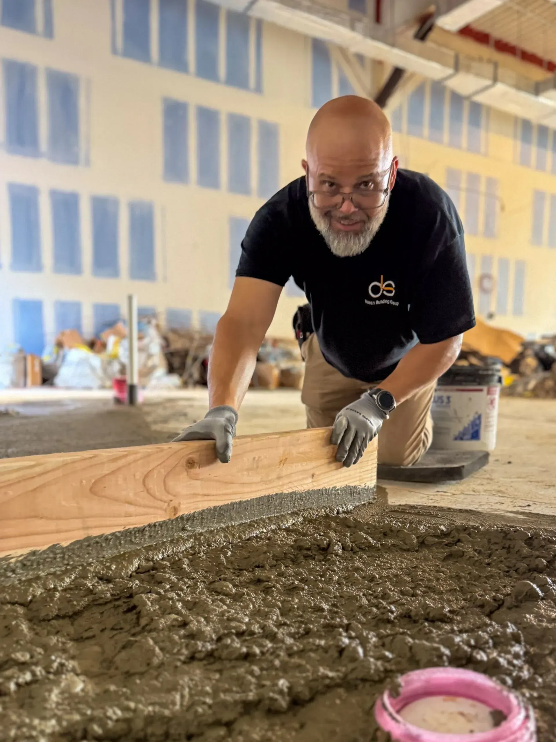 Man leveling wet cement with a wooden plank in an unfinished building. He wears a black shirt, gloves, and has a beard.