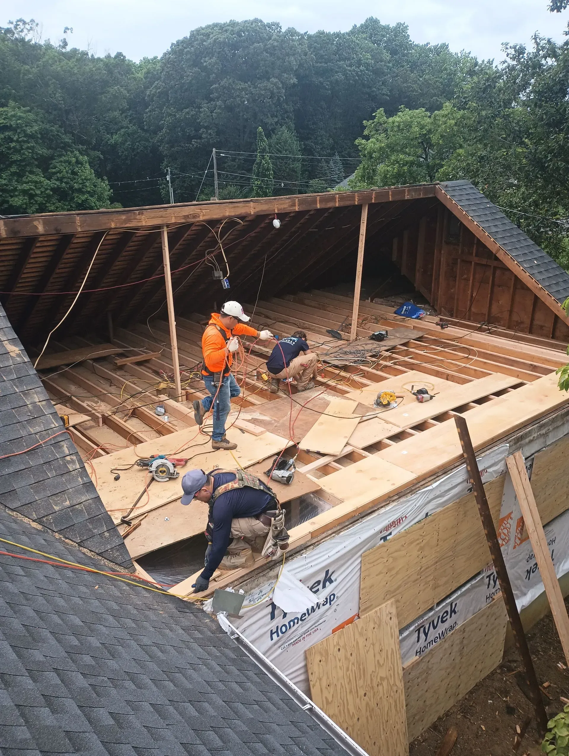 Construction workers rebuilding a roof:  plywood, tools,  in an attic. One wears orange, another blue. Trees in the background.
