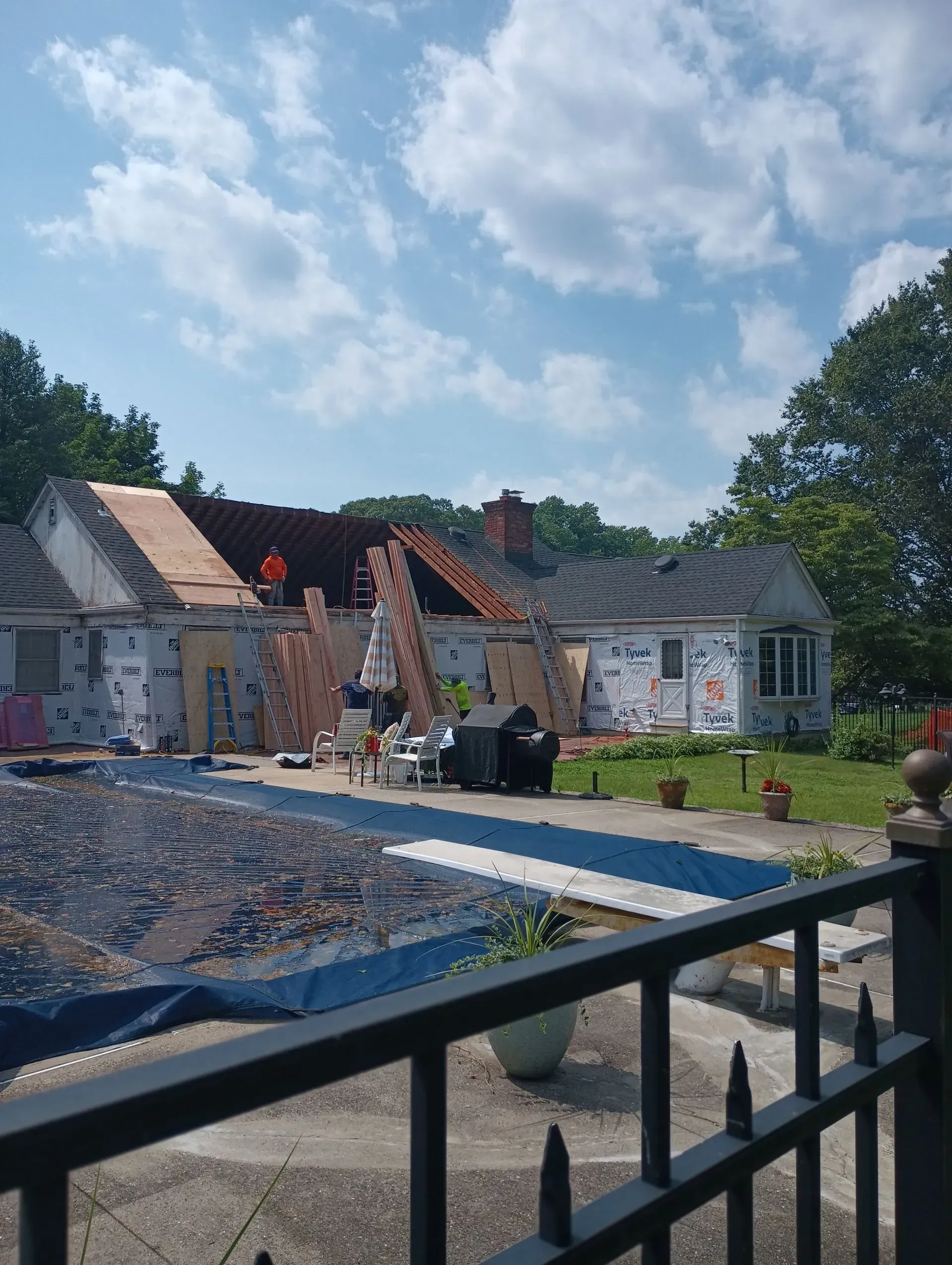 House under roof repair; blue sky with clouds, construction workers on roof, covered pool in foreground.