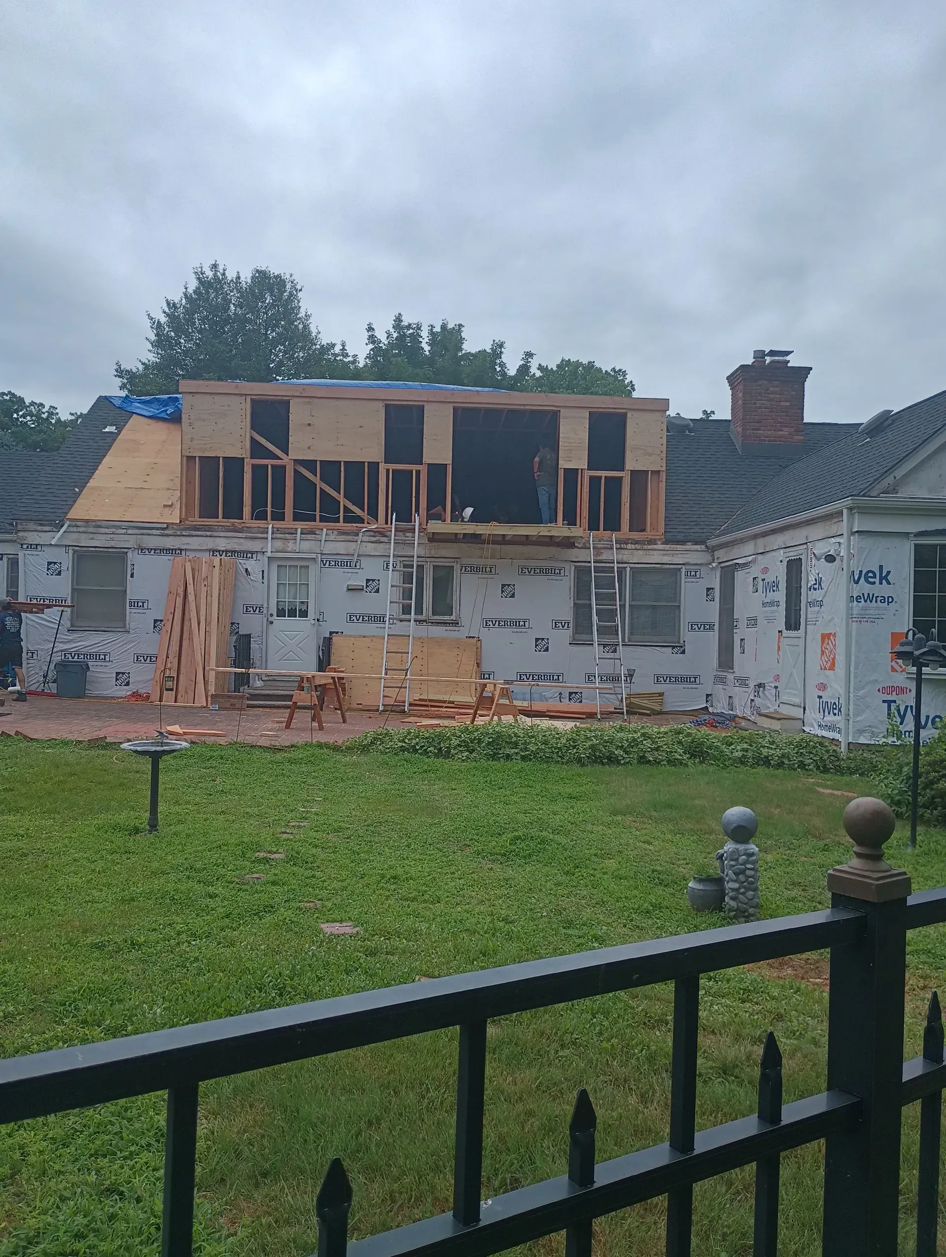 House under construction, addition being built on the roof, siding and wood frame visible. Green grass and cloudy sky.