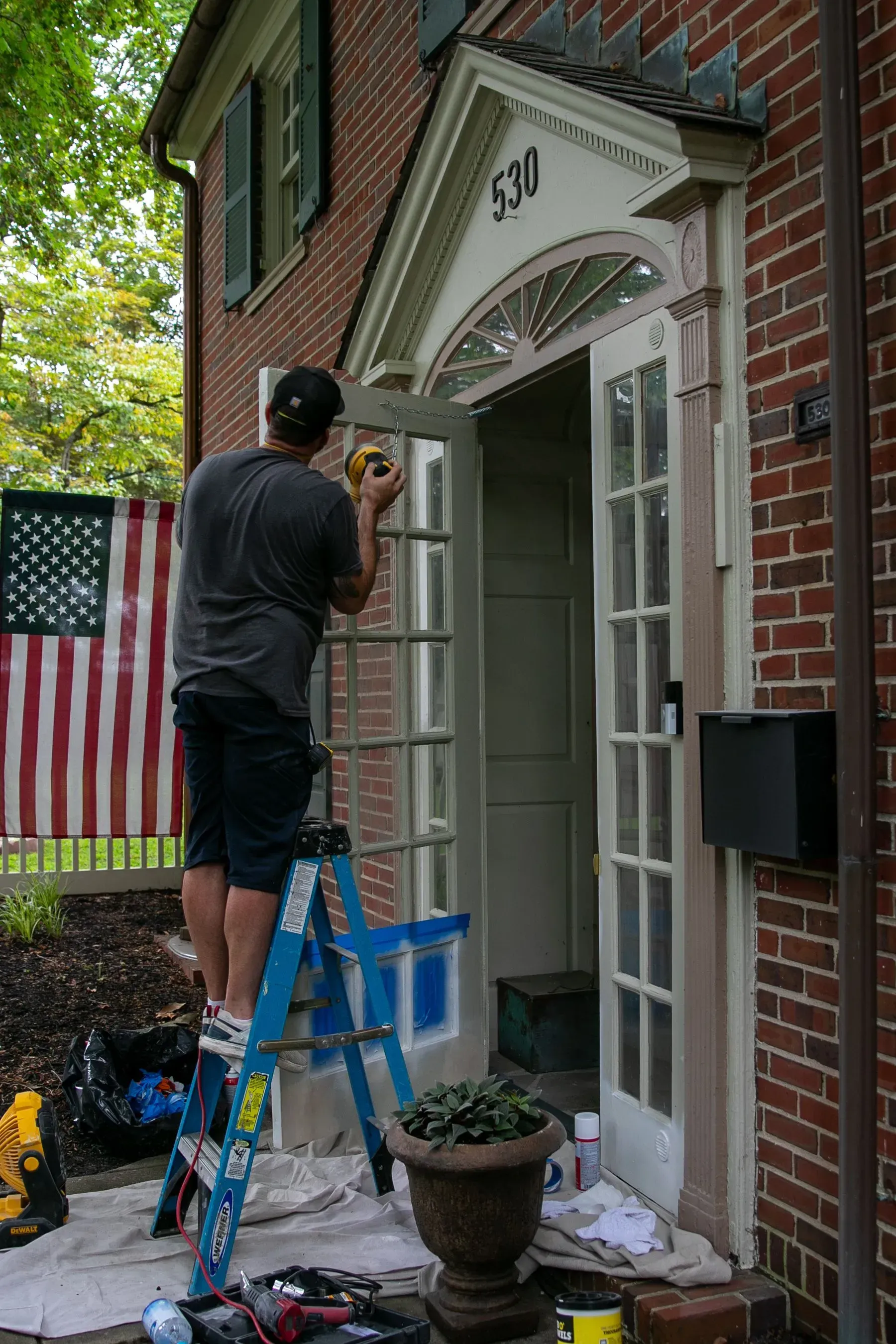 Man on a ladder painting the doorway of a brick house; an American flag hangs nearby.
