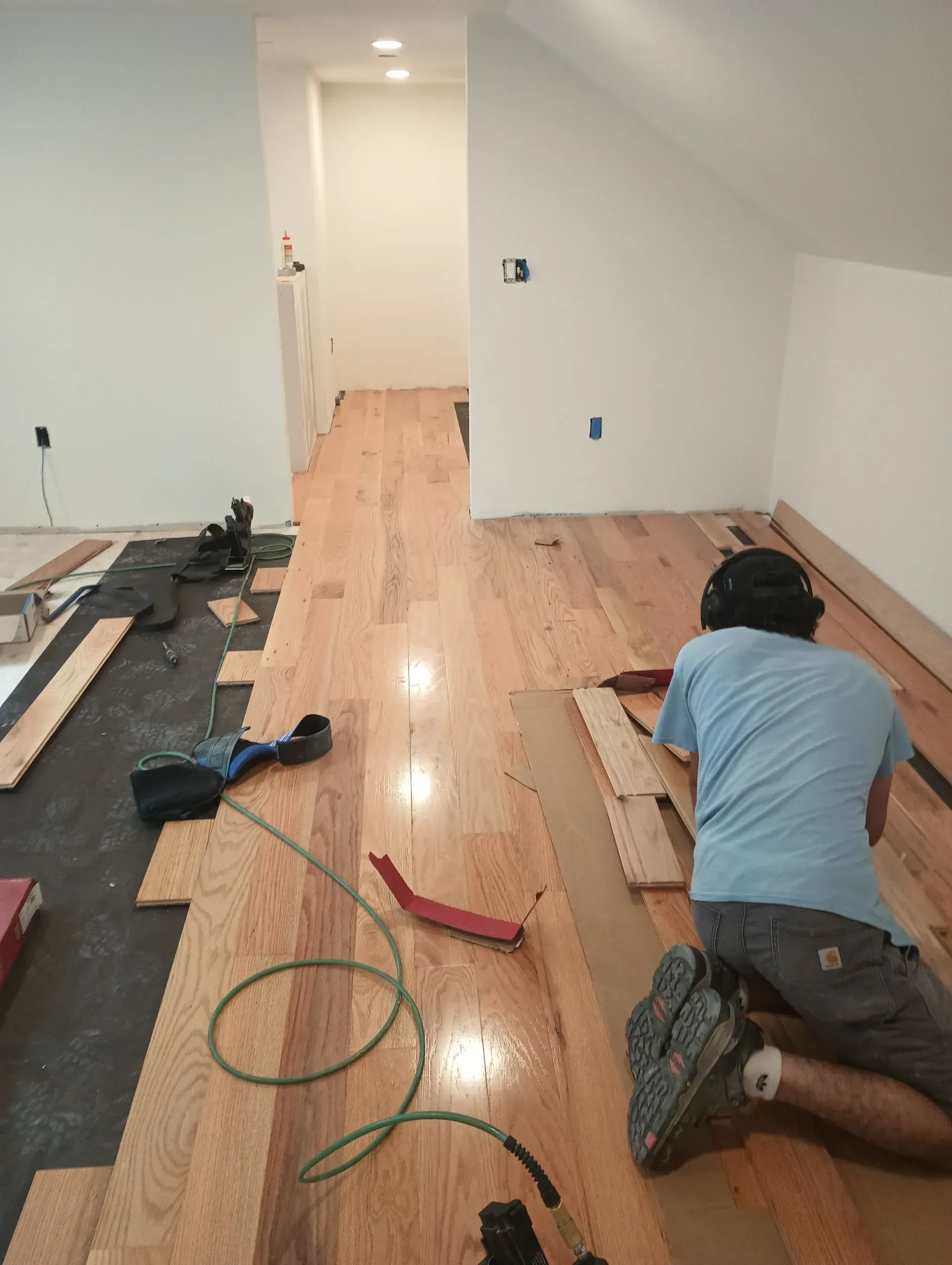 Man installing hardwood floors in a room with white walls.  He's kneeling, using tools, with unfinished flooring in the foreground.