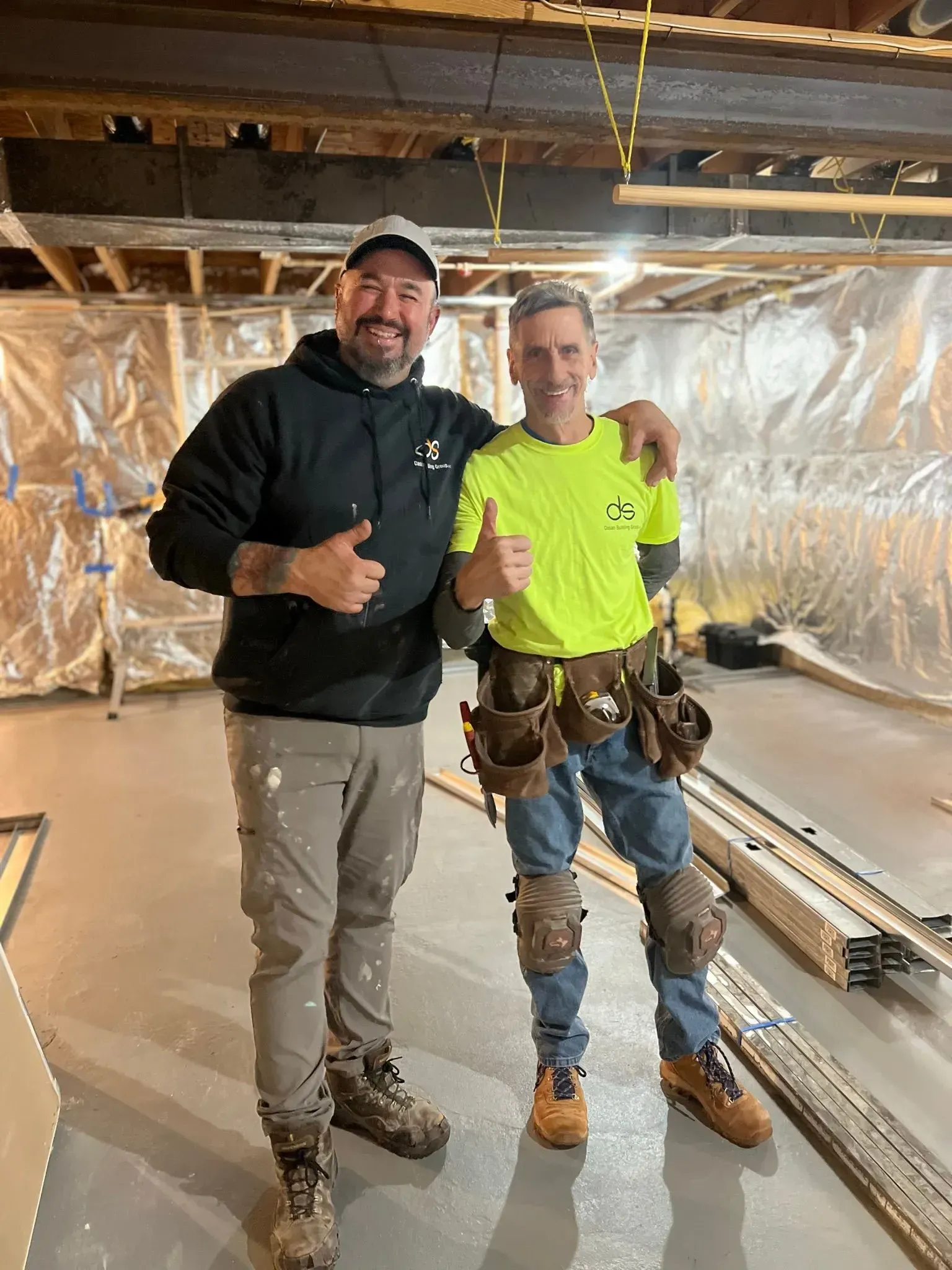 Two construction workers smiling and giving thumbs up in a basement.