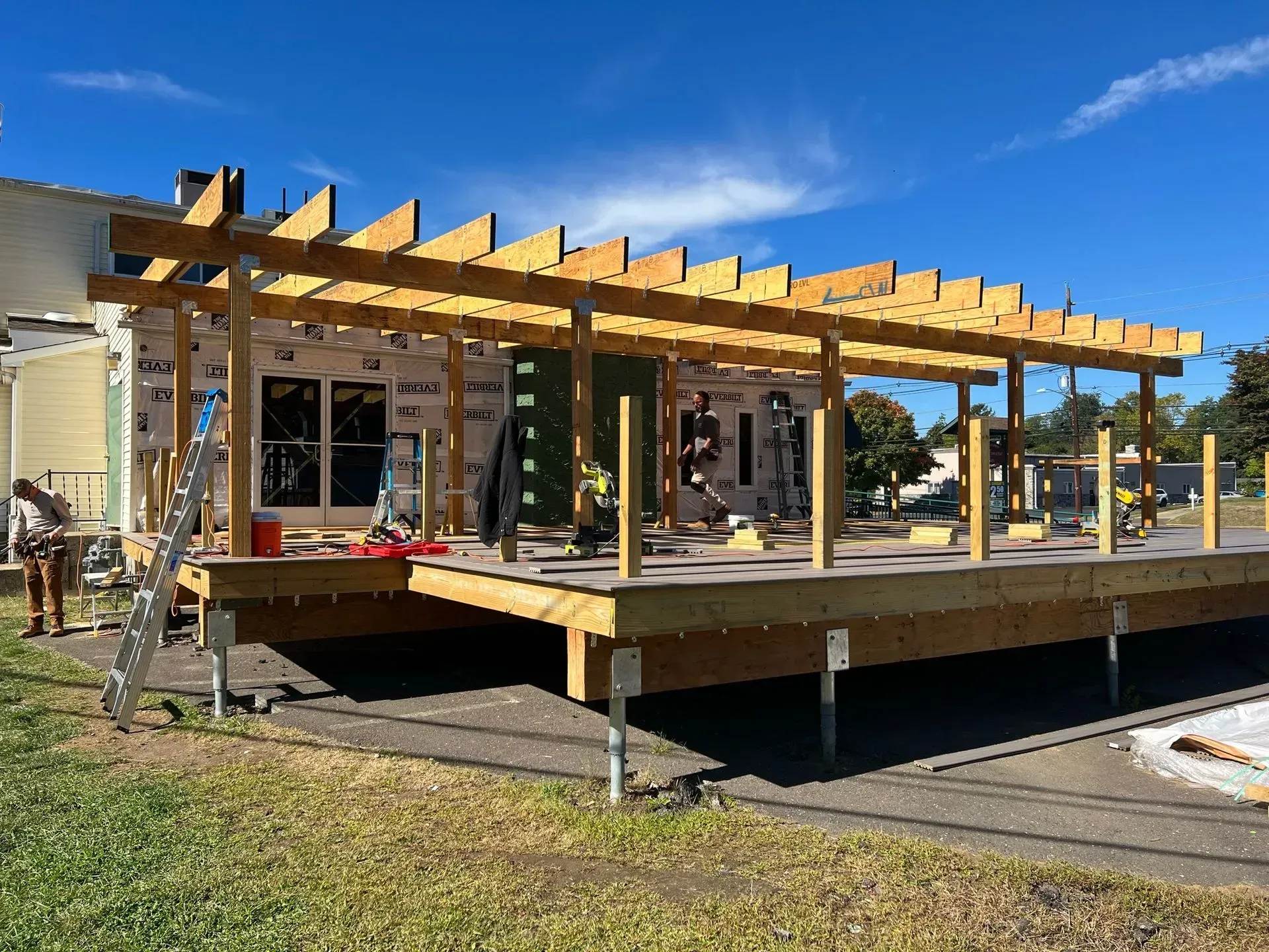 Construction of a wooden deck with a pergola attached to a building under a blue sky.