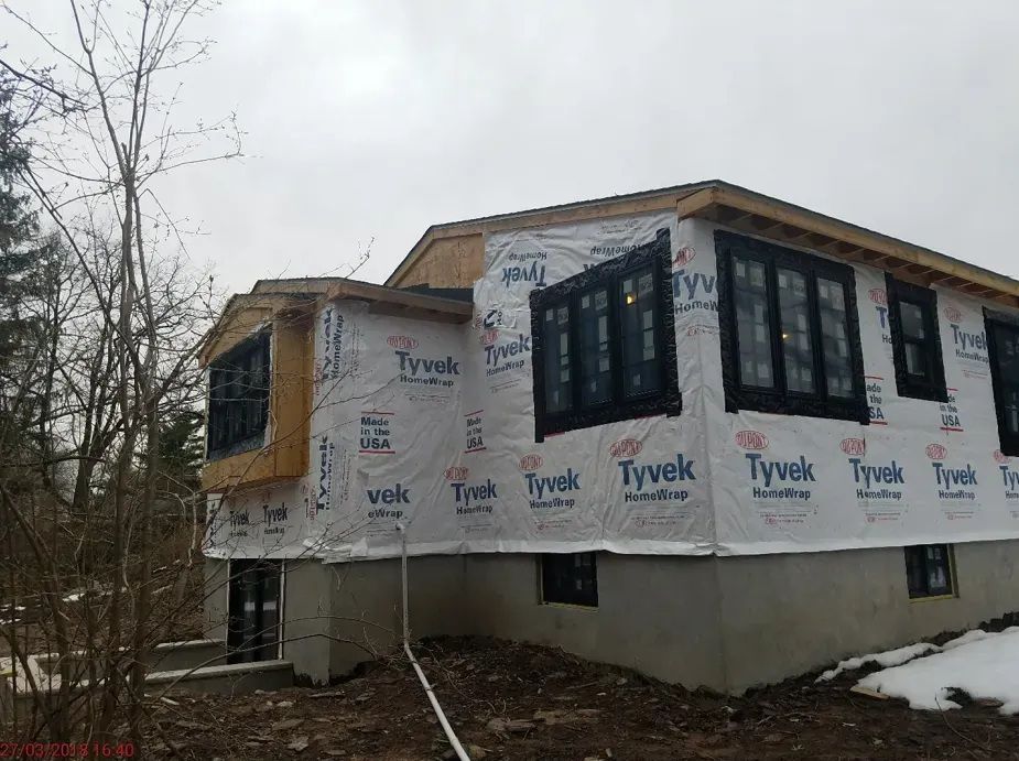 Partially built house wrapped in Tyvek, with large black-framed windows and bare trees nearby