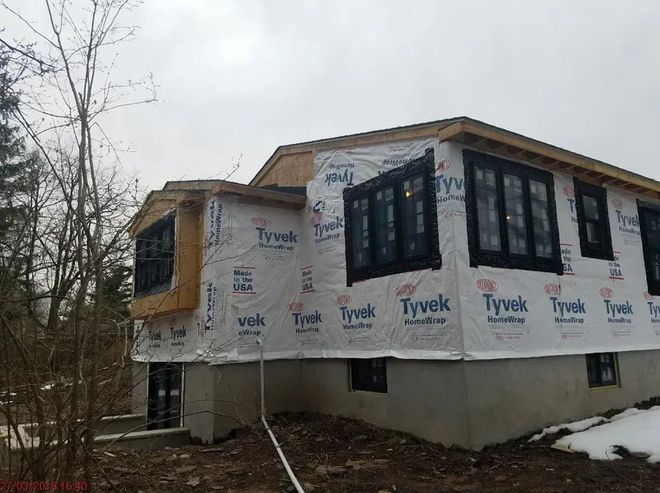 Partially built house wrapped in Tyvek, with large black-framed windows and bare trees nearby