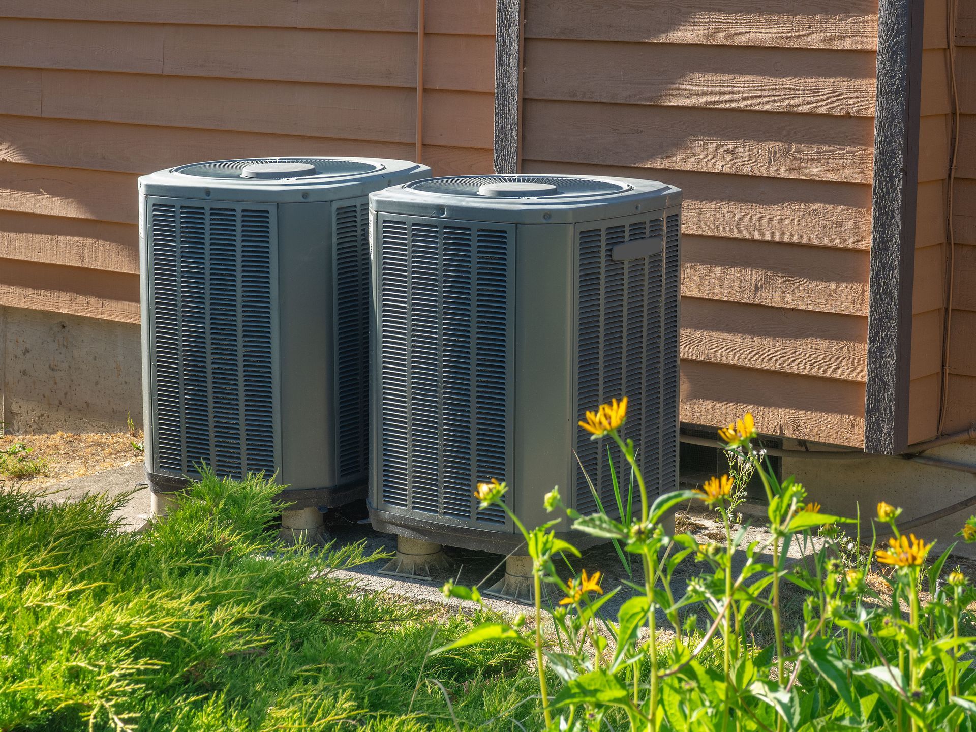 Two air conditioning units next to a wooden building, with grass and flowers in front.