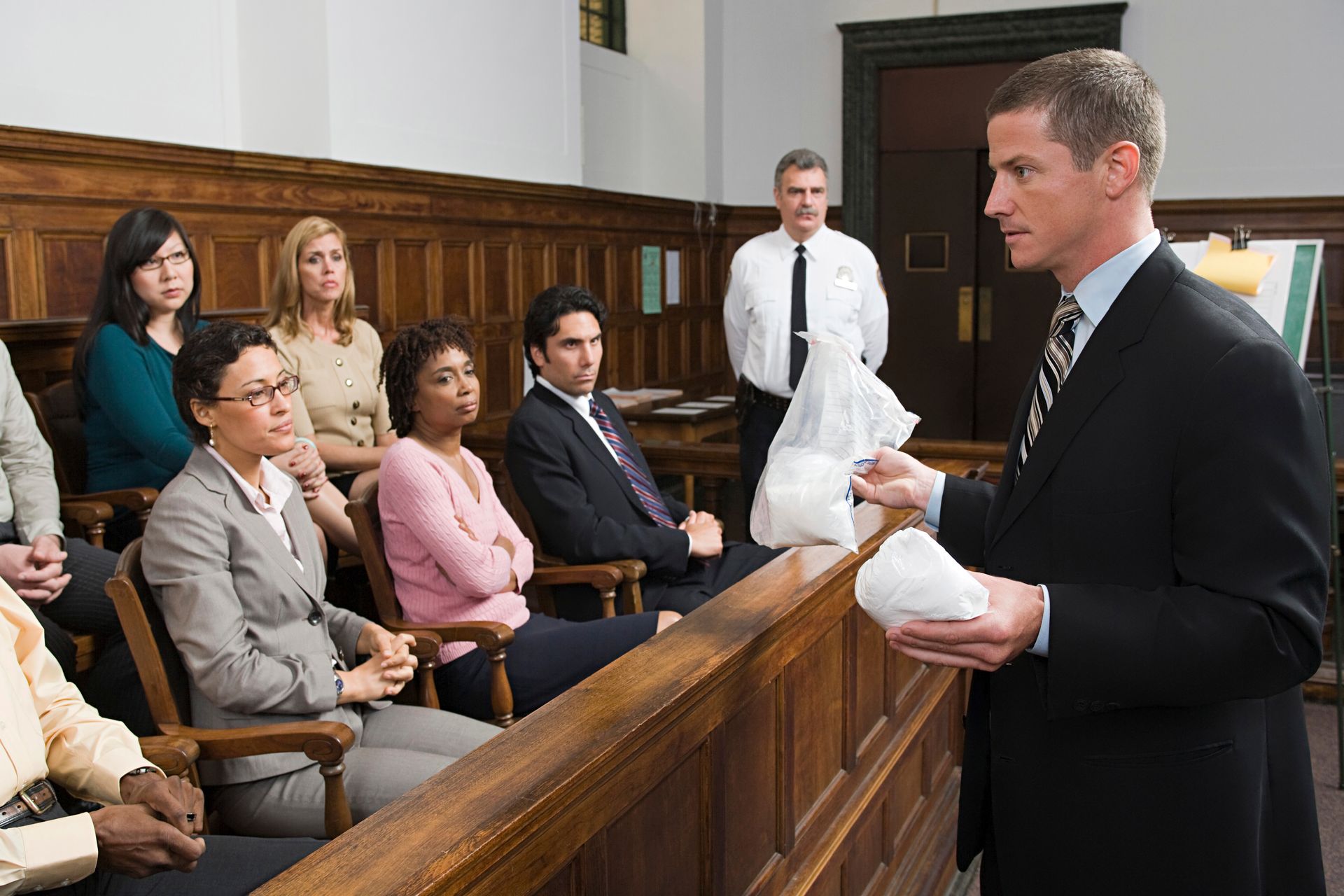 Attorney addressing jury in a courtroom while holding a wrapped item; seated jurors listen attentively.