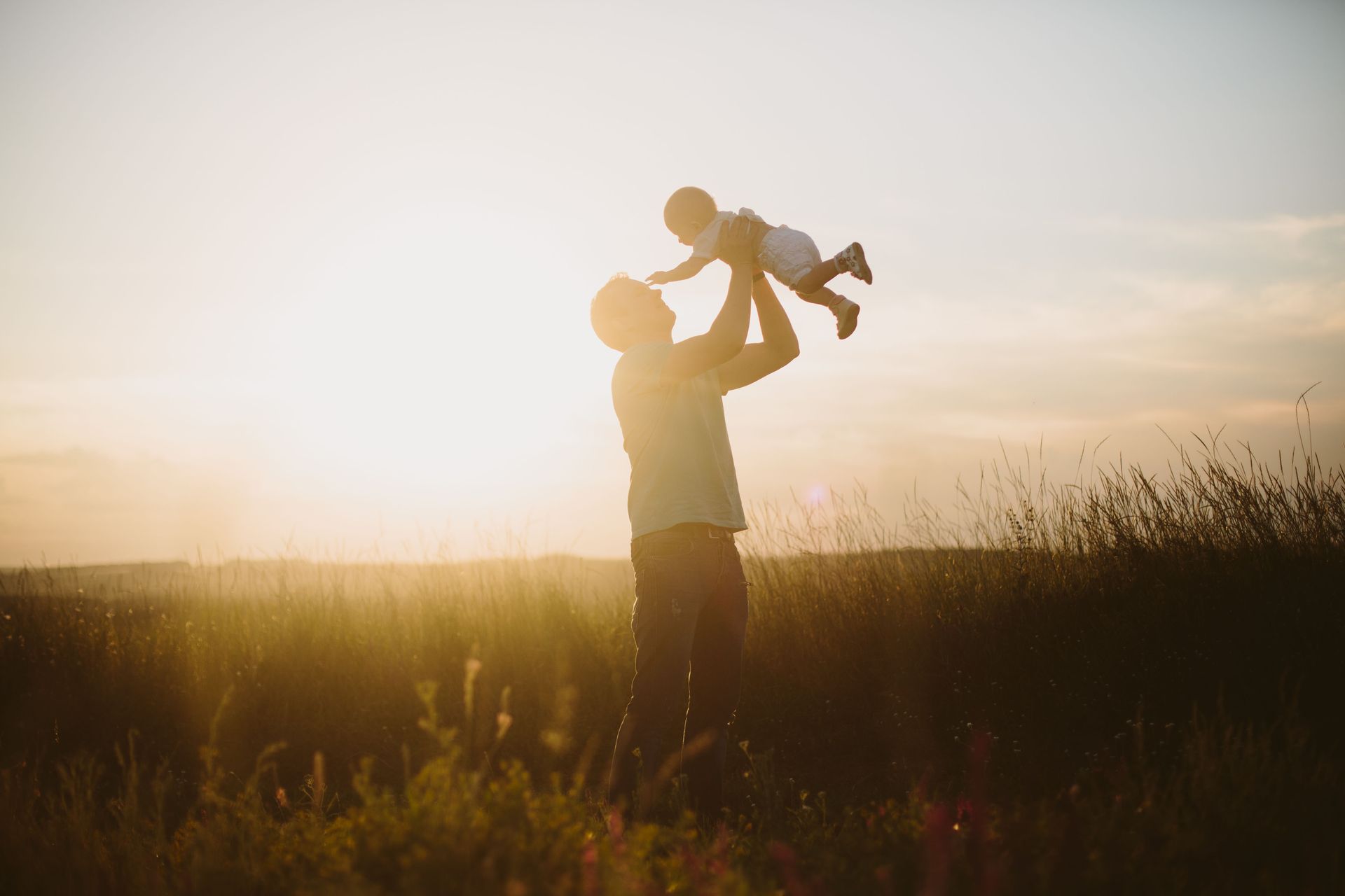 Silhouetted adult lifting a child overhead in a grassy field at sunset