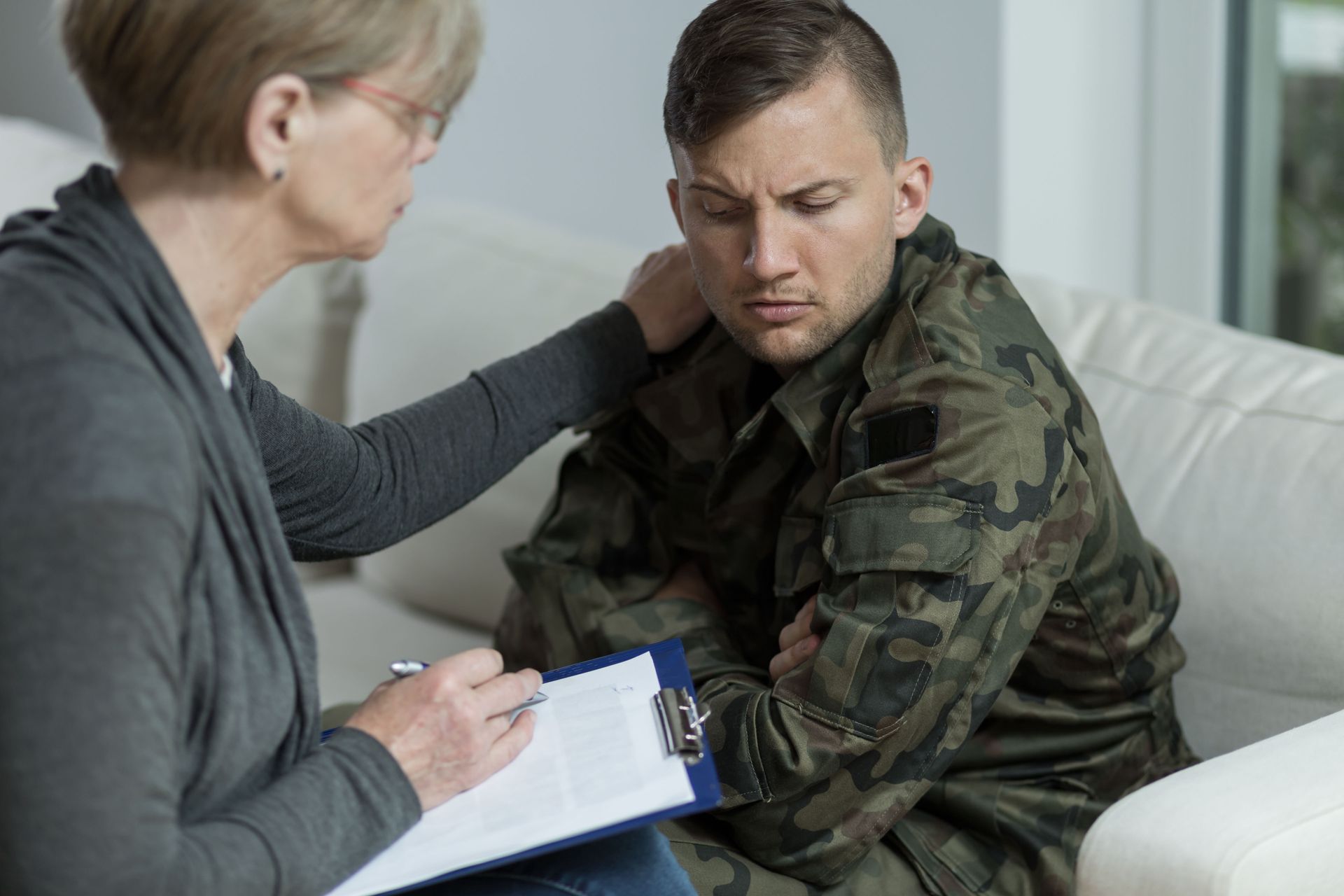 Counselor comfortingly speaks with a distressed soldier on a couch, clipboard in hand.