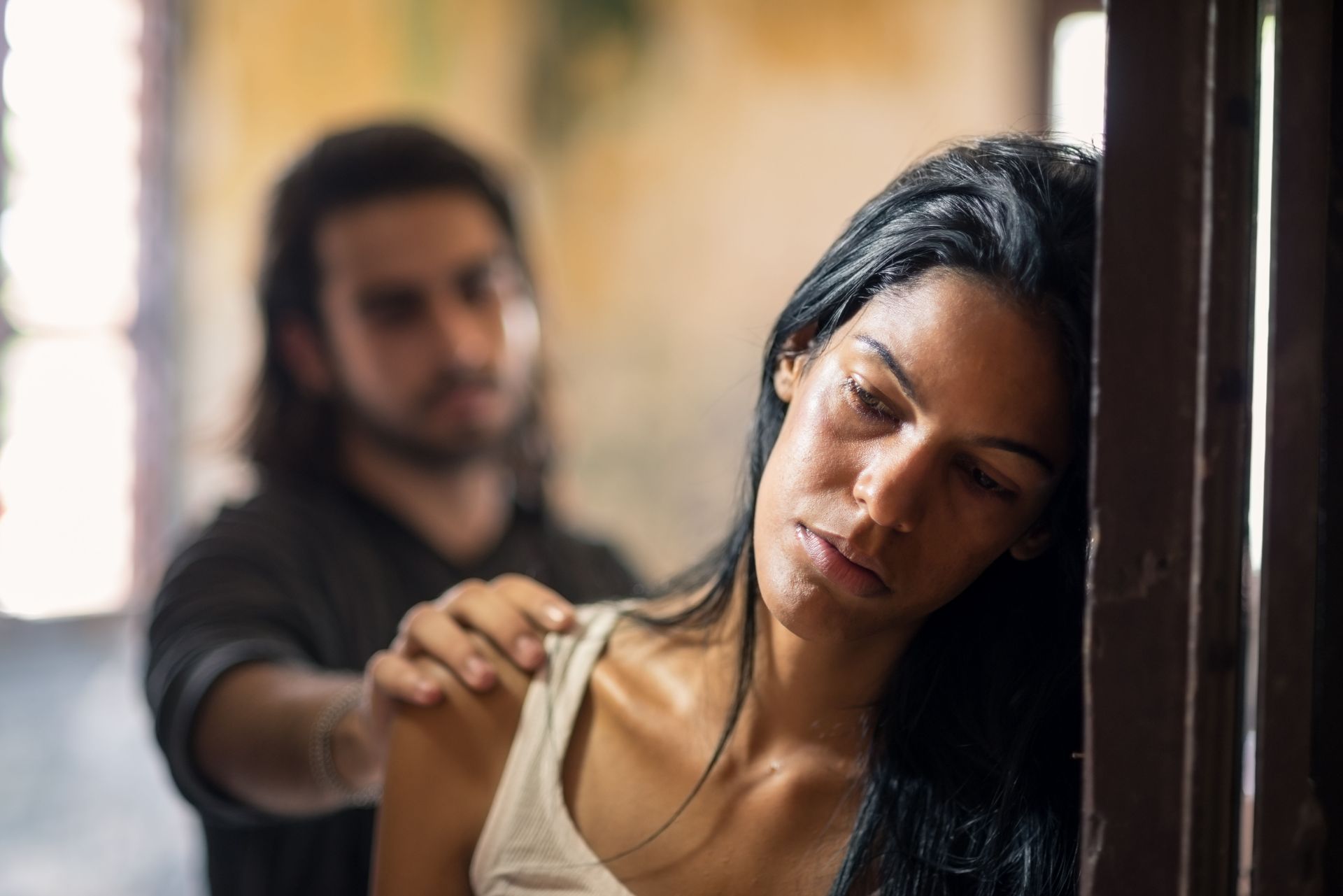 Woman leans against a doorway while a man reaches toward her shoulder in a dim, tense indoor scene