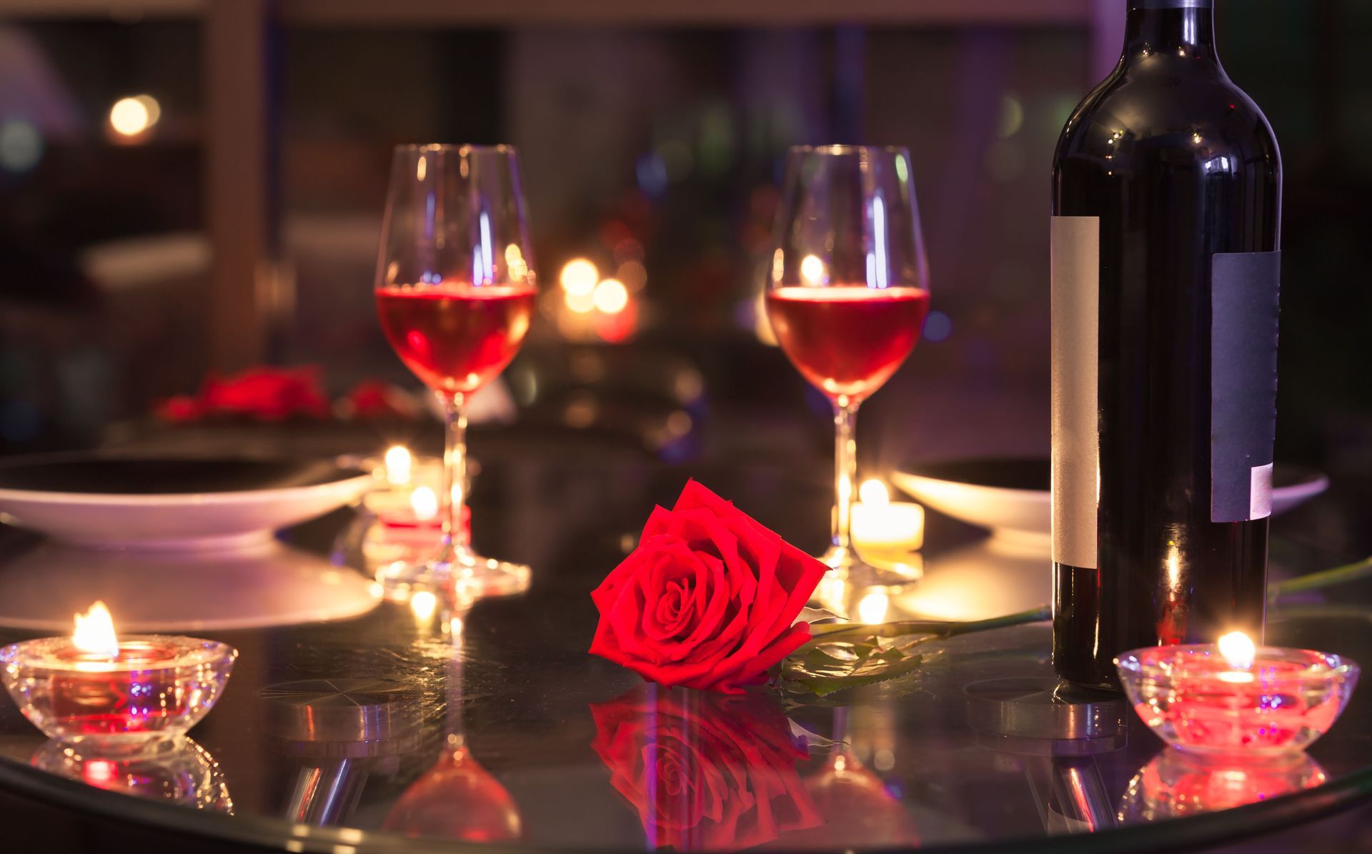 Romantic candlelit table with red wine, roses, and a bottle on a reflective surface.