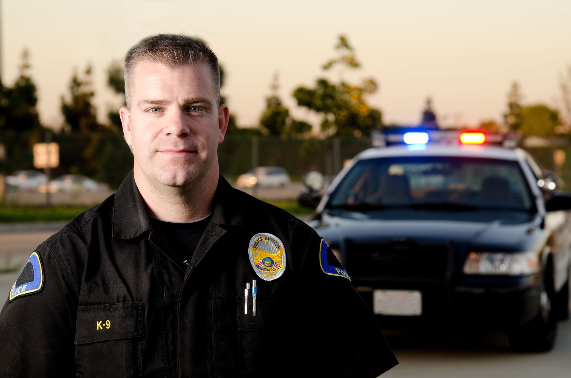 Police officer standing in front of a patrol car with flashing lights at dusk