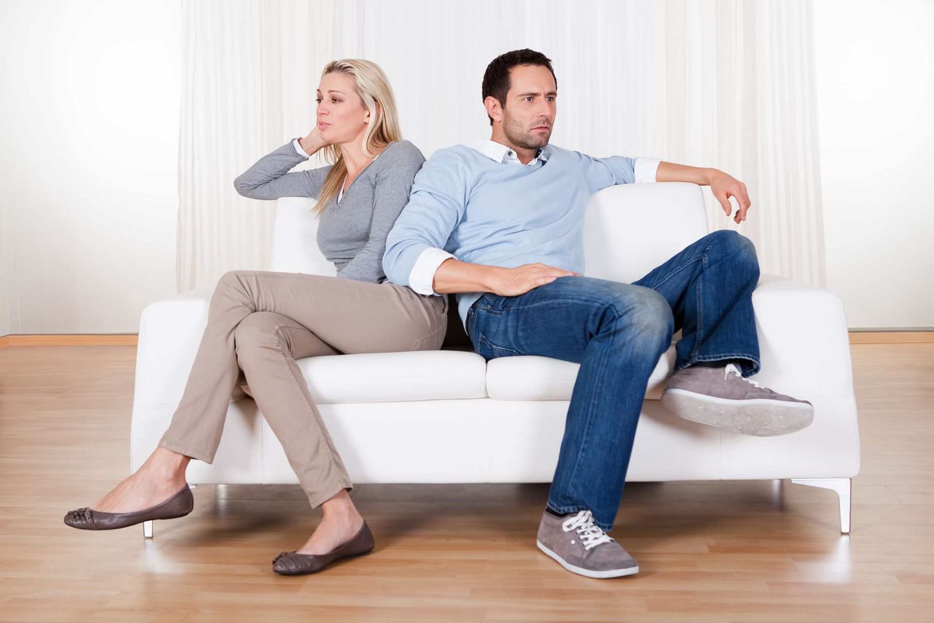 Two people sitting back-to-back on a white couch, looking away in a bright living room.