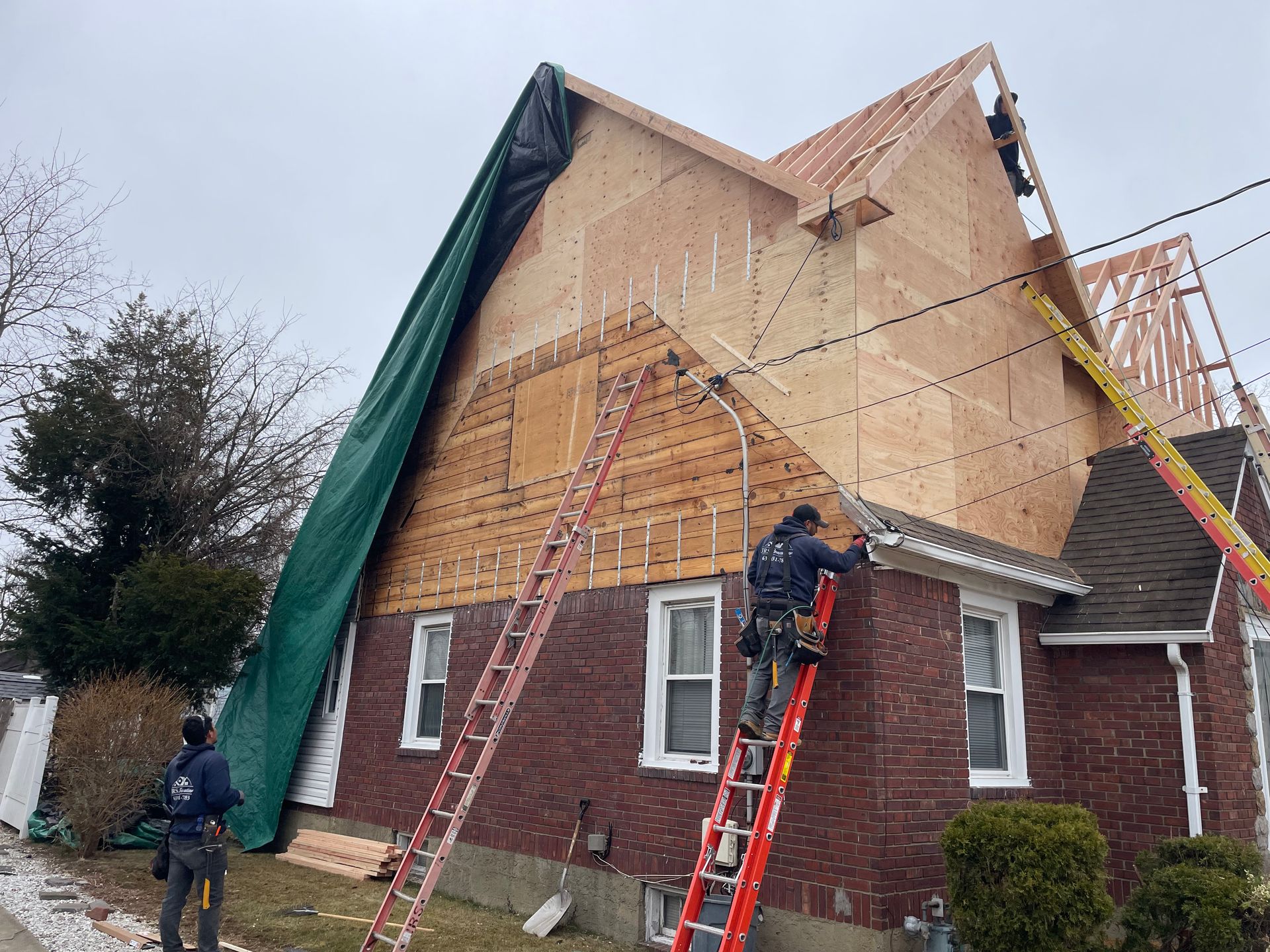 Construction workers building a house roof; two on ladders, plywood walls, green tarp, red brick building, cloudy day.
