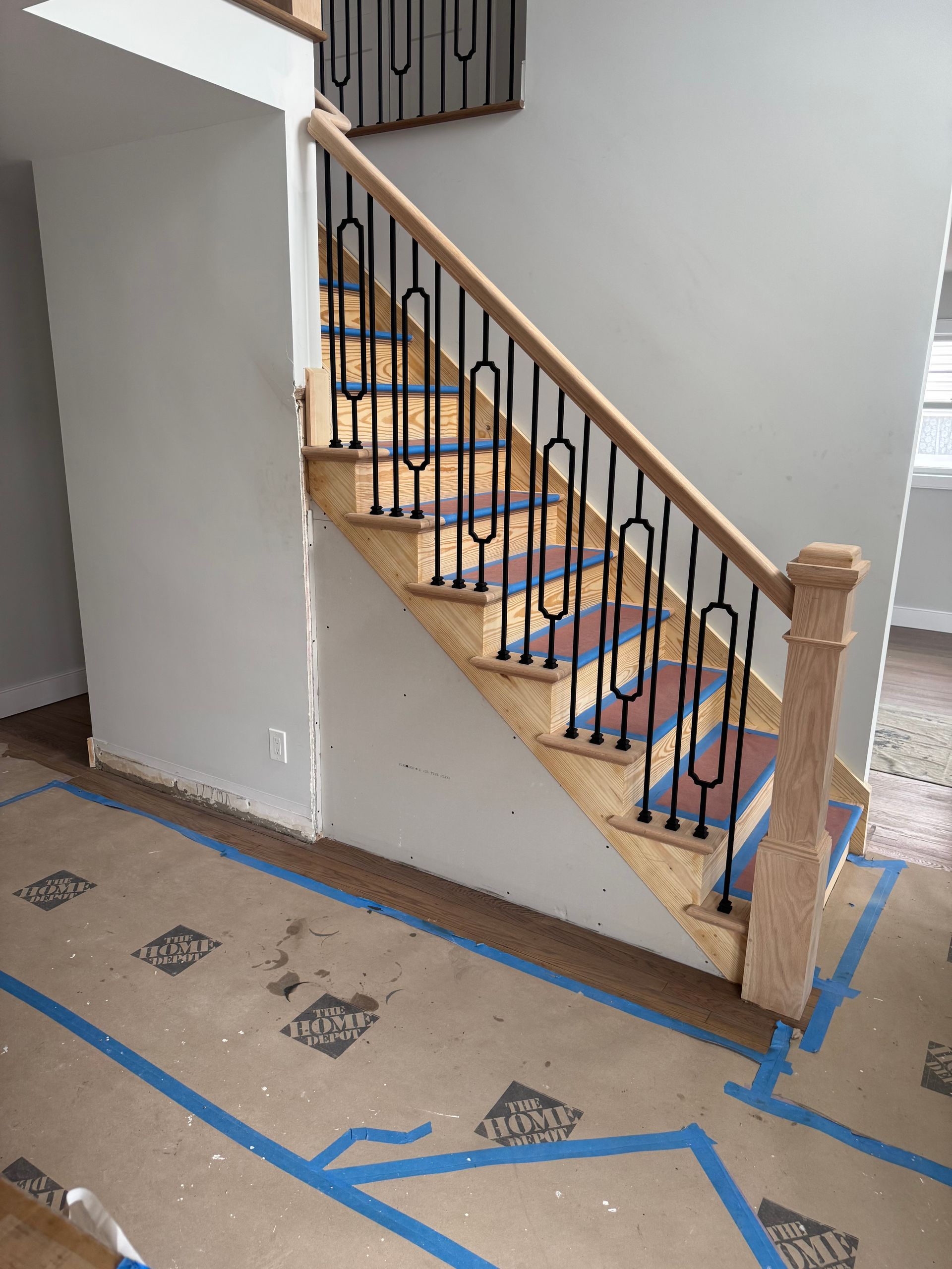Staircase with light wood steps, black metal balusters, and a wooden handrail. Blue tape lines the floor.