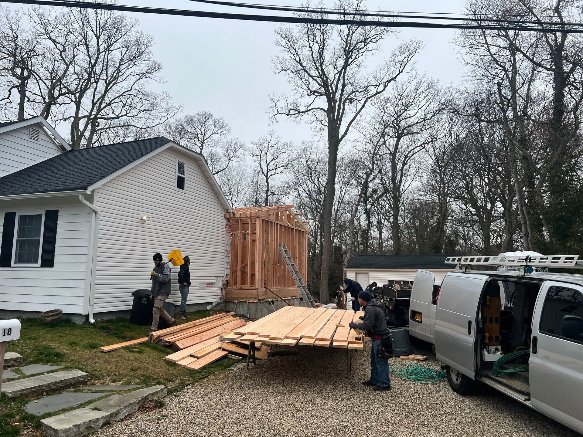 Construction workers building an addition to a white house. Lumber and a van are present. Overcast day.