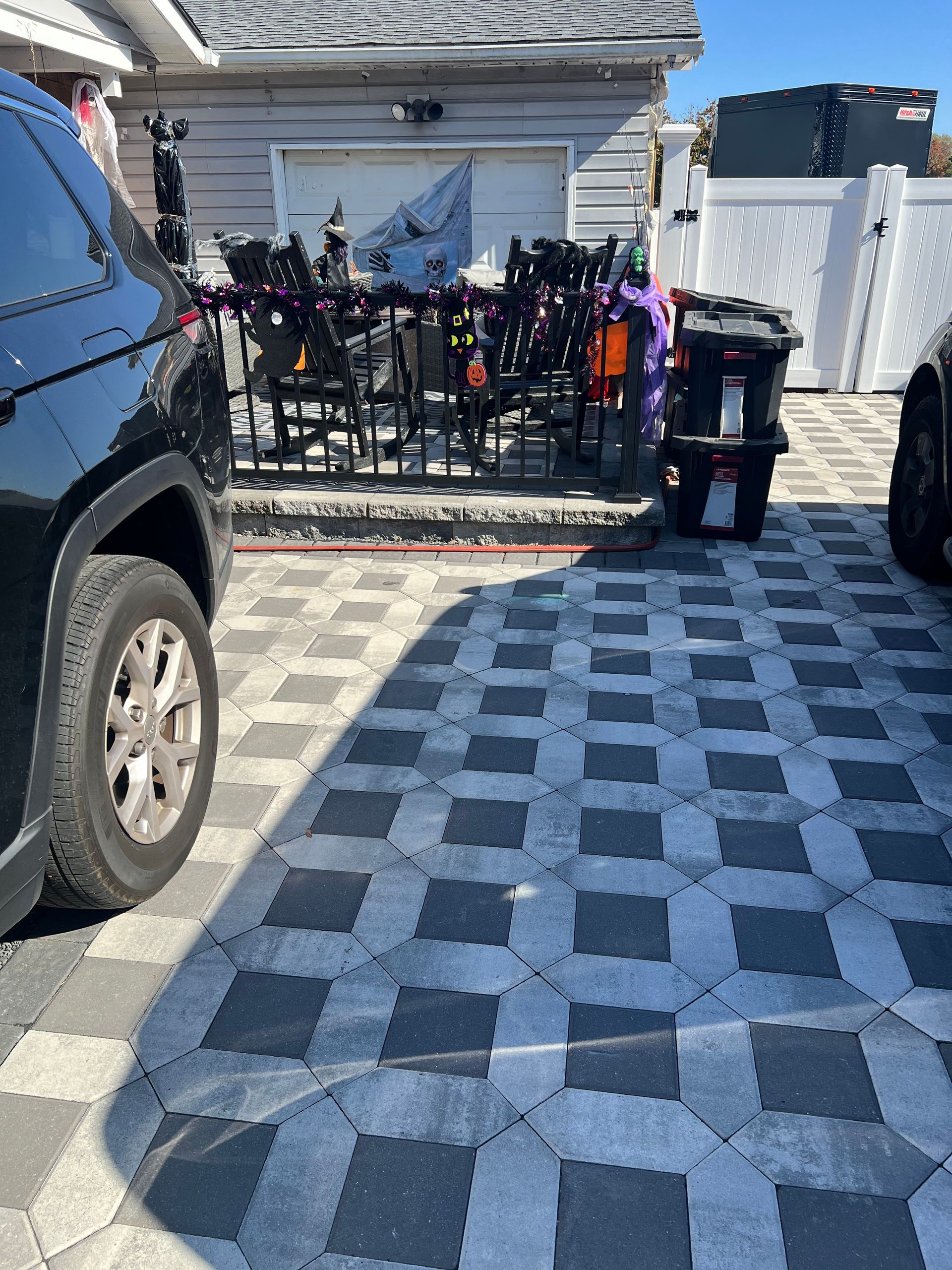 Driveway paved with checkered stones, black car on left, trash cans on right, fence and bikes in background.