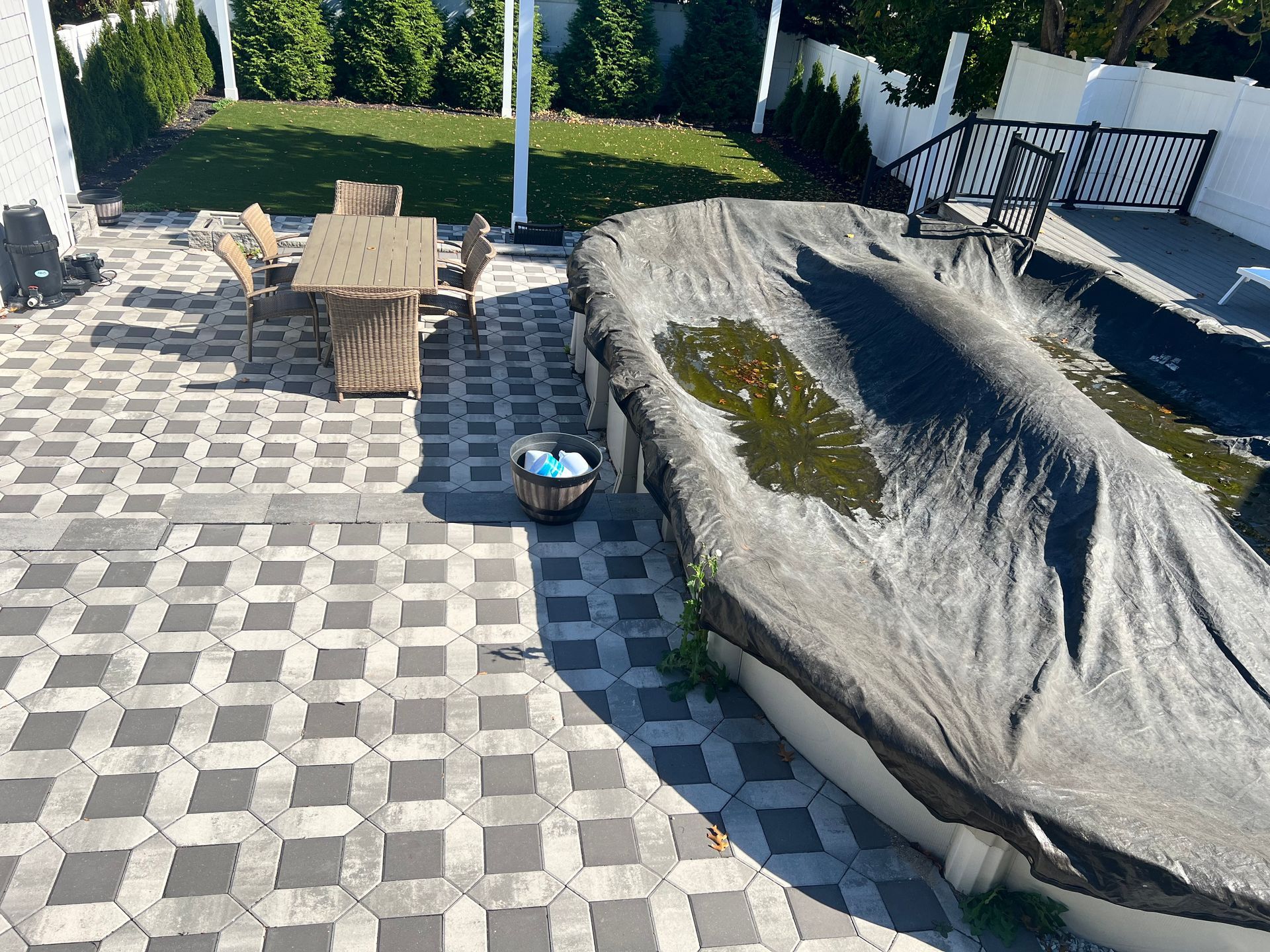 Patio with gray patterned paving, covered pool, and wooden patio furniture. Green grass and white fence visible.