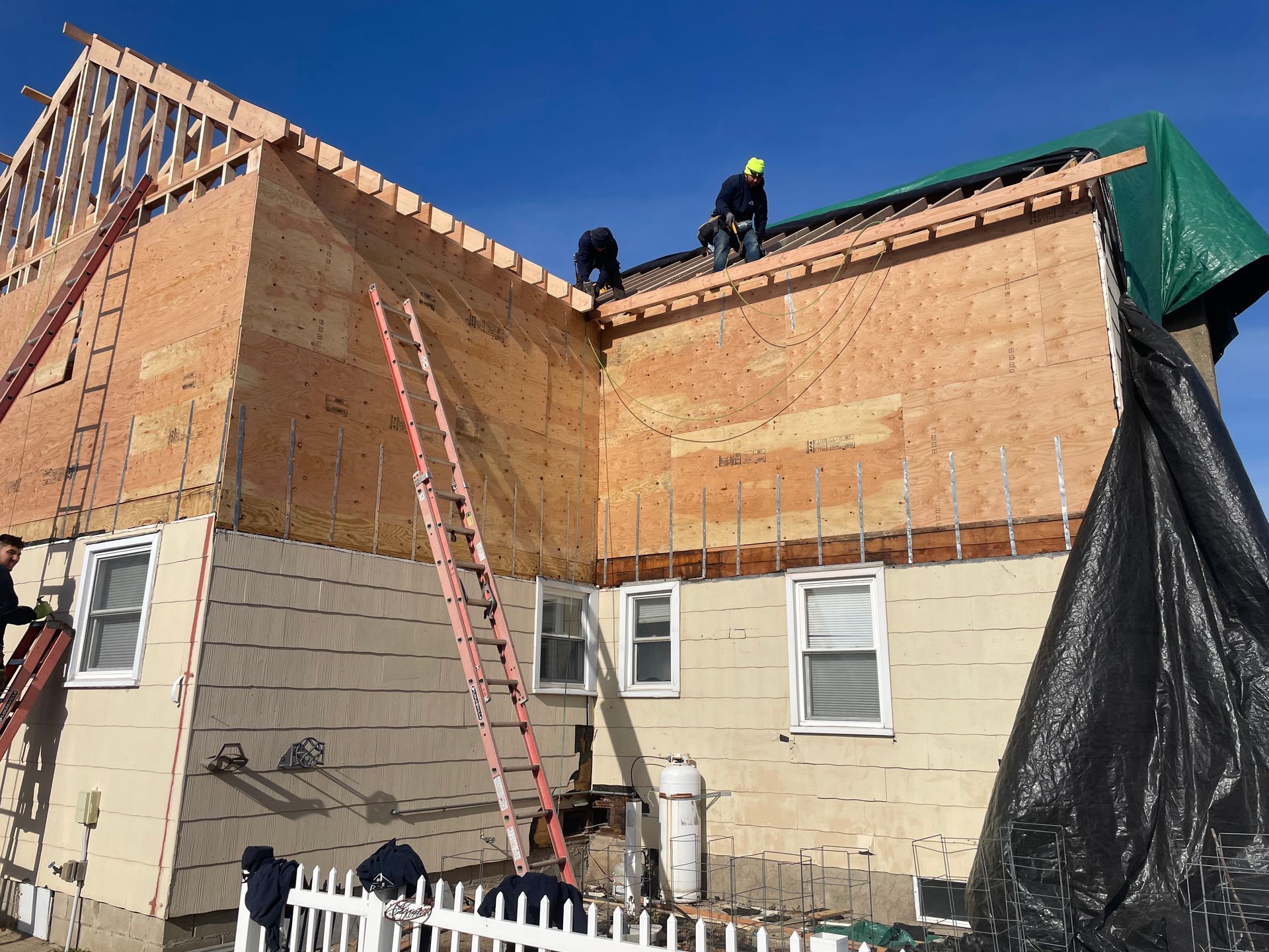 Construction workers on a house, adding a roof. Wooden frame and plywood visible. Ladders and blue sky.