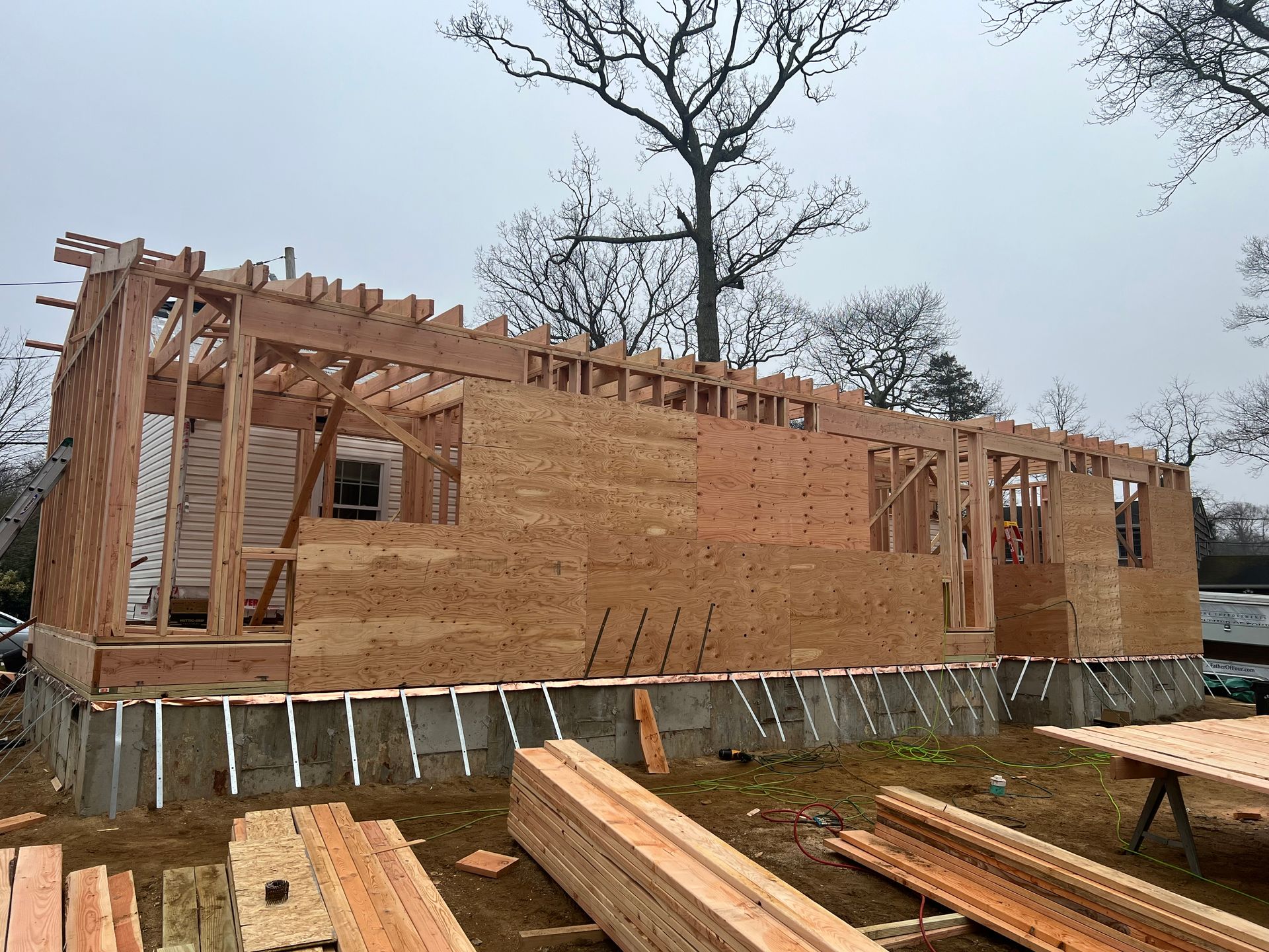 Wood-framed house under construction; exposed studs and plywood walls; lumber pile in foreground.