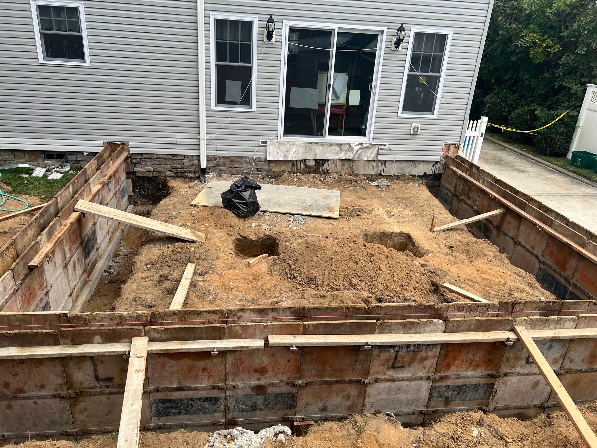 Construction site: Excavated rectangular area with wooden forms, dirt, and house in the background.