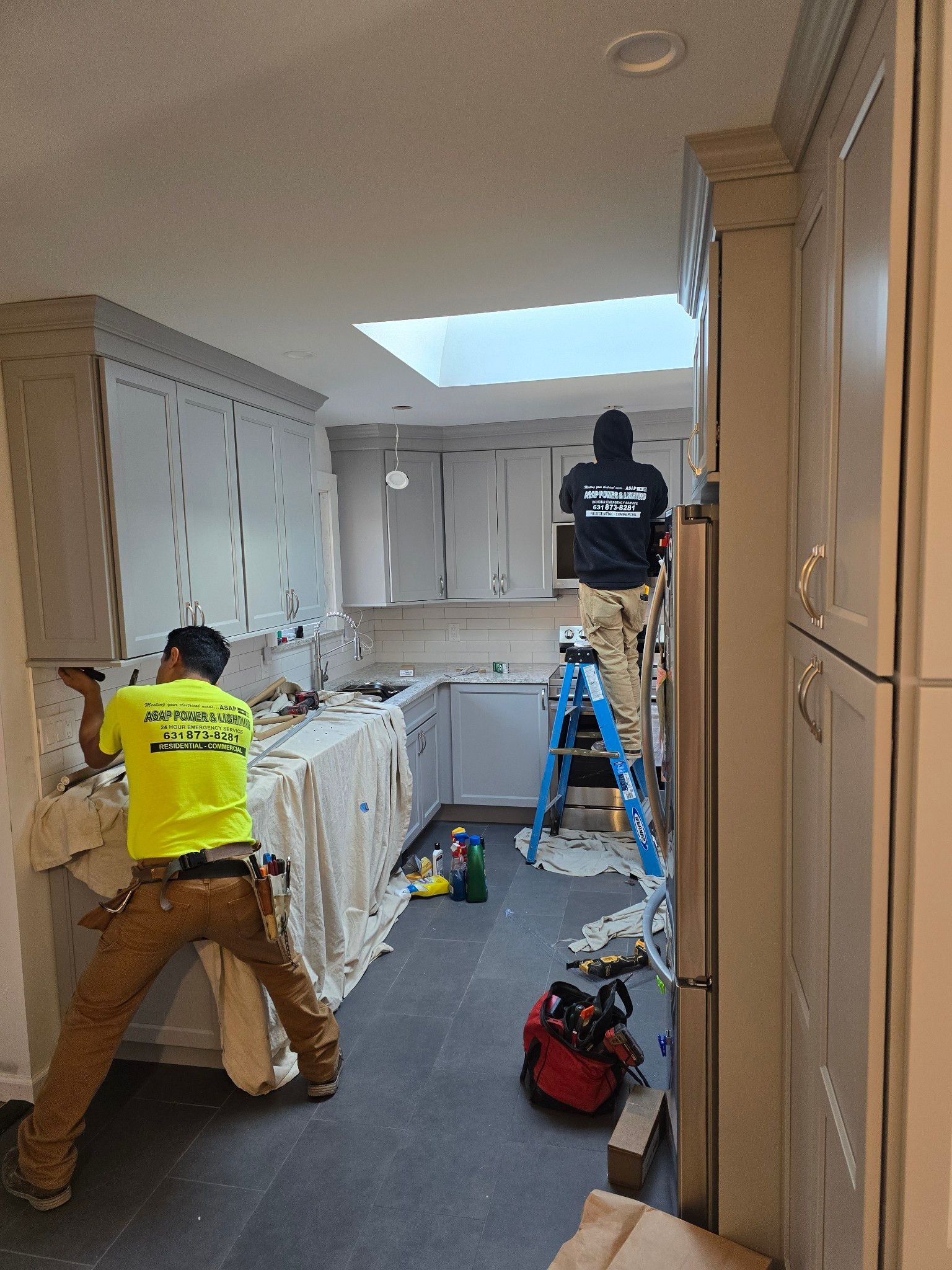 Two workers installing kitchen tiles, cabinets visible. One on a ladder, another tiling near the counter.