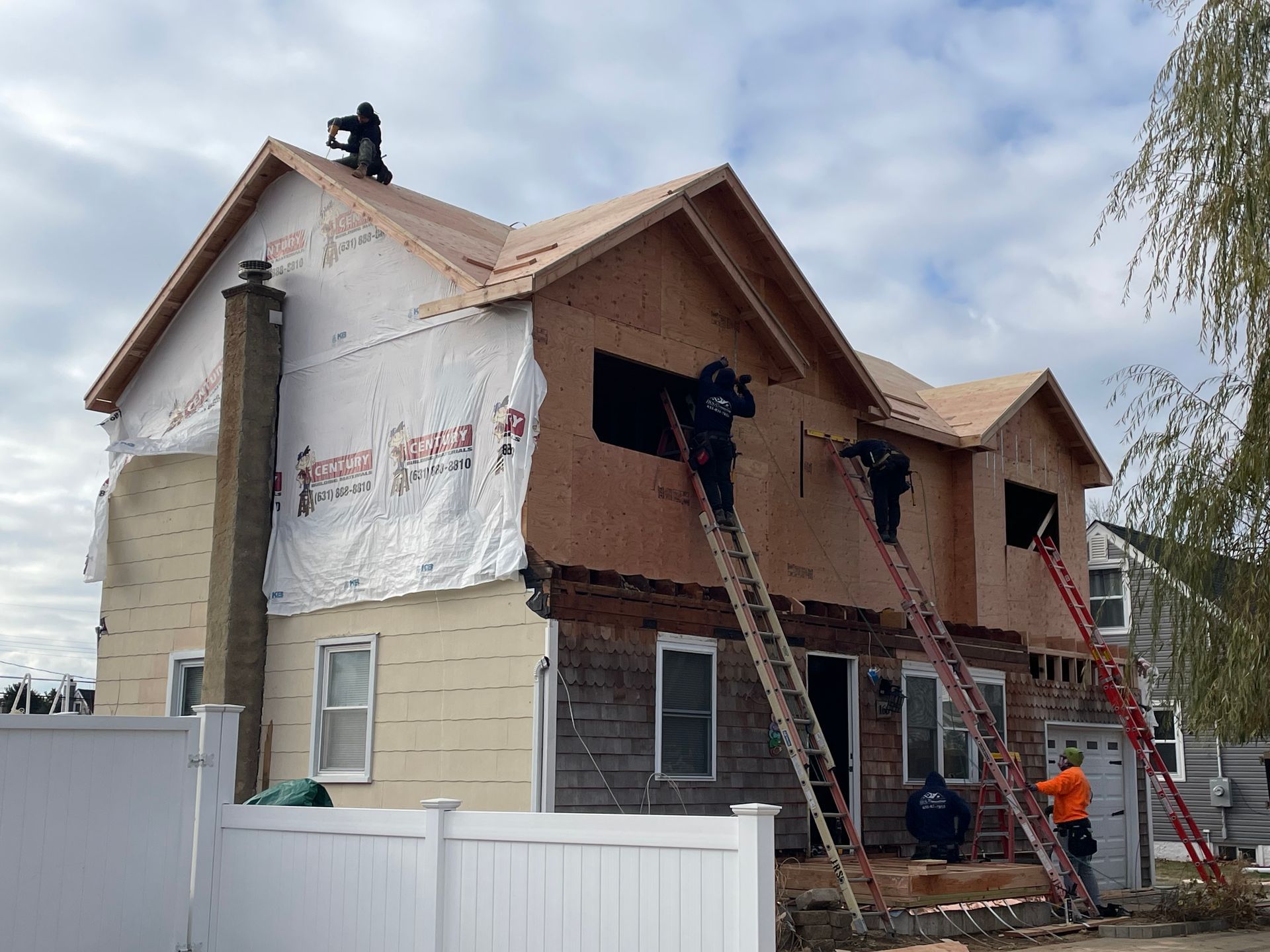 Construction workers on ladders working on a two-story house. Partially sided with a chimney and new roof.