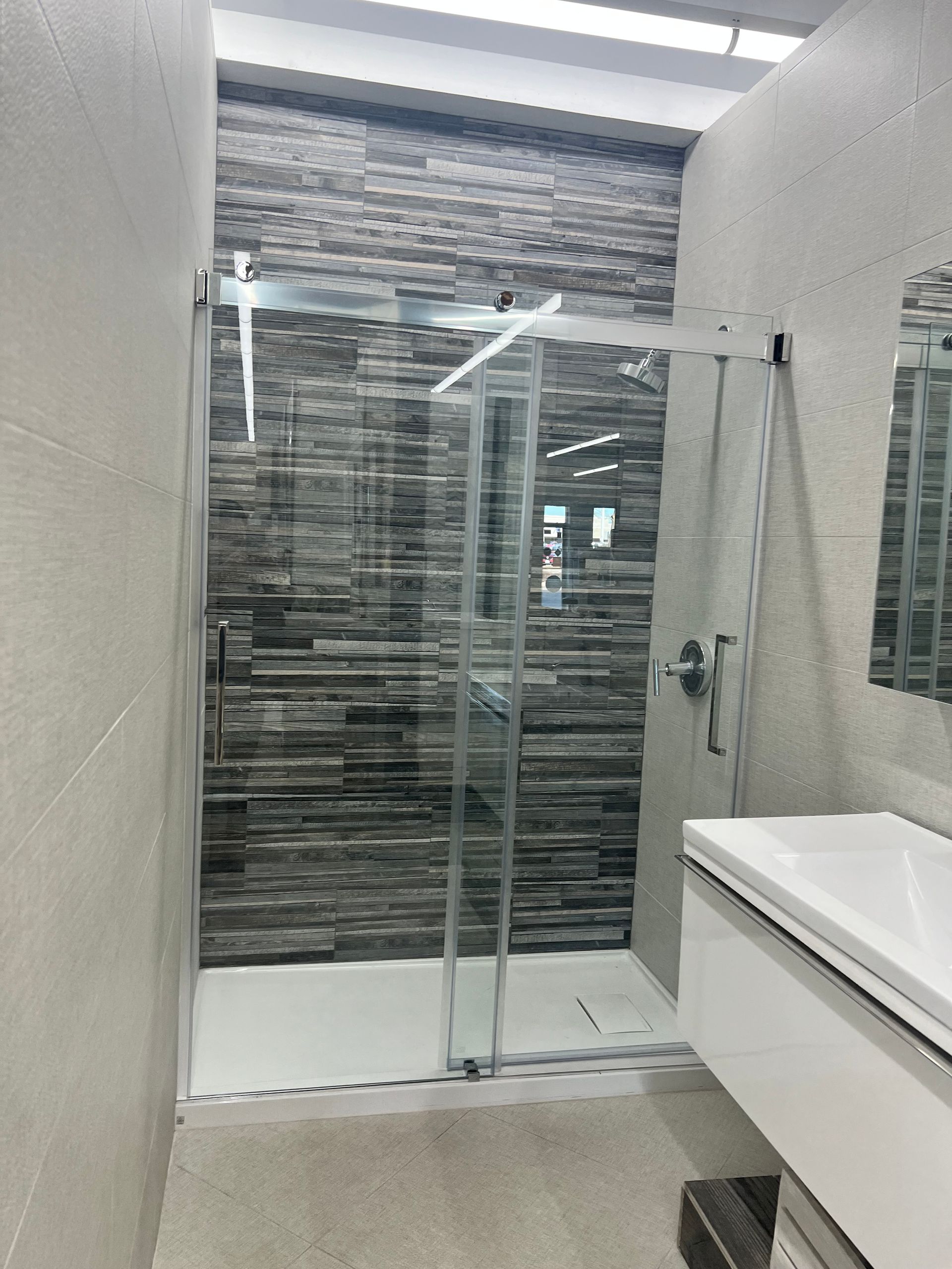 Modern bathroom with a glass shower, textured gray tile wall, white sink, and light gray walls.