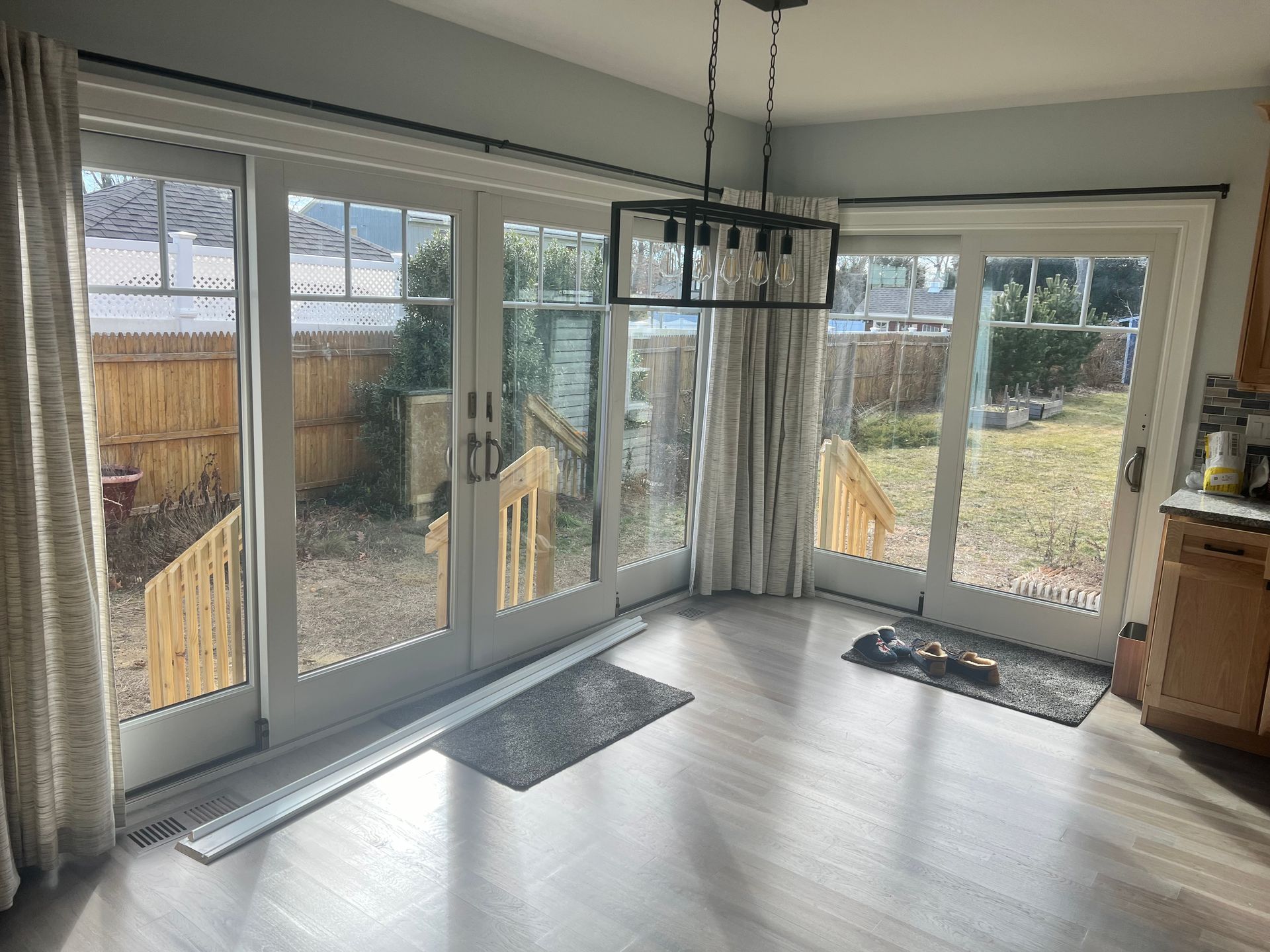 Kitchen with sliding glass doors overlooking a backyard. Light-colored curtains, hardwood floors, and a hanging light fixture.