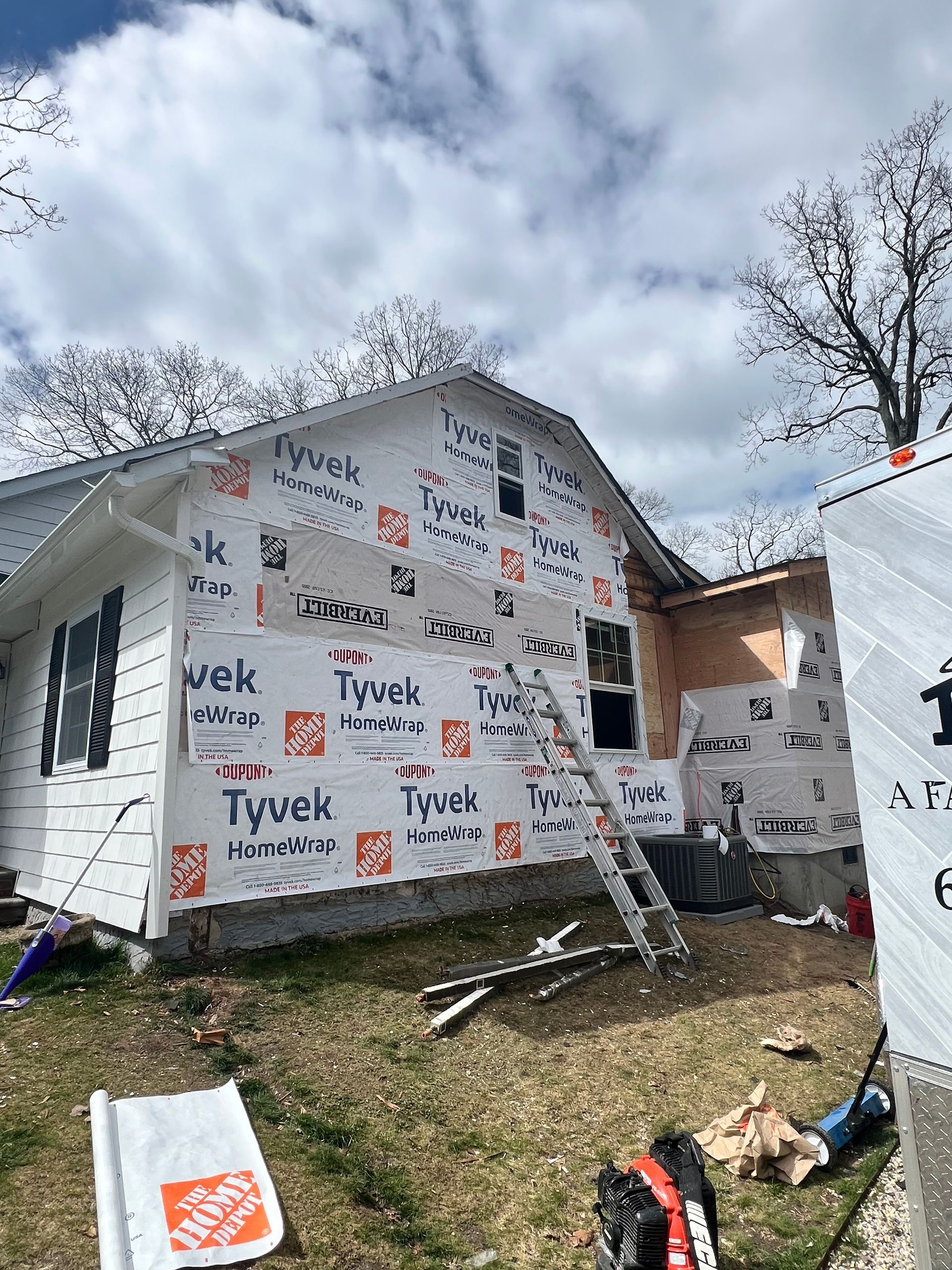 House exterior under construction, covered in Tyvek wrap. White siding, windows, and ladder visible. Cloudy sky.